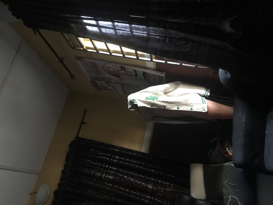 The image depicts an indoor scene within a room located in Oba-Ile, Nigeria. A standing human figure is positioned centrally, partially obscured by shadows, wearing an open, cream-colored varsity-style jacket with green trim, green buttons, and a prominent green "LA" emblem on the left chest. The figure's head is not visible, appearing to be covered or obscured by the jacket's hood, and bare legs of light skin tone are visible below the jacket.
The room features light brown/tan walls, a white ceiling with a visible beam, and a white decorative light fixture in the upper left corner. Two windows are present; the left is mostly covered by a dark, checkered curtain, while the right window, partially covered by a matching curtain, reveals bright exterior light filtered through vertical metal grilles. Dark curtain rods with finials are mounted above both windows. Two light-colored posters are affixed to the wall between the windows. Furniture includes a black armchair or sofa in the left foreground with visible wear and a white armrest, and a dark-colored sofa armrest in the right foreground. The room is dimly lit, with strong backlighting from the right window creating deep shadows. No discernible actions or interactions beyond the figure standing are visible.