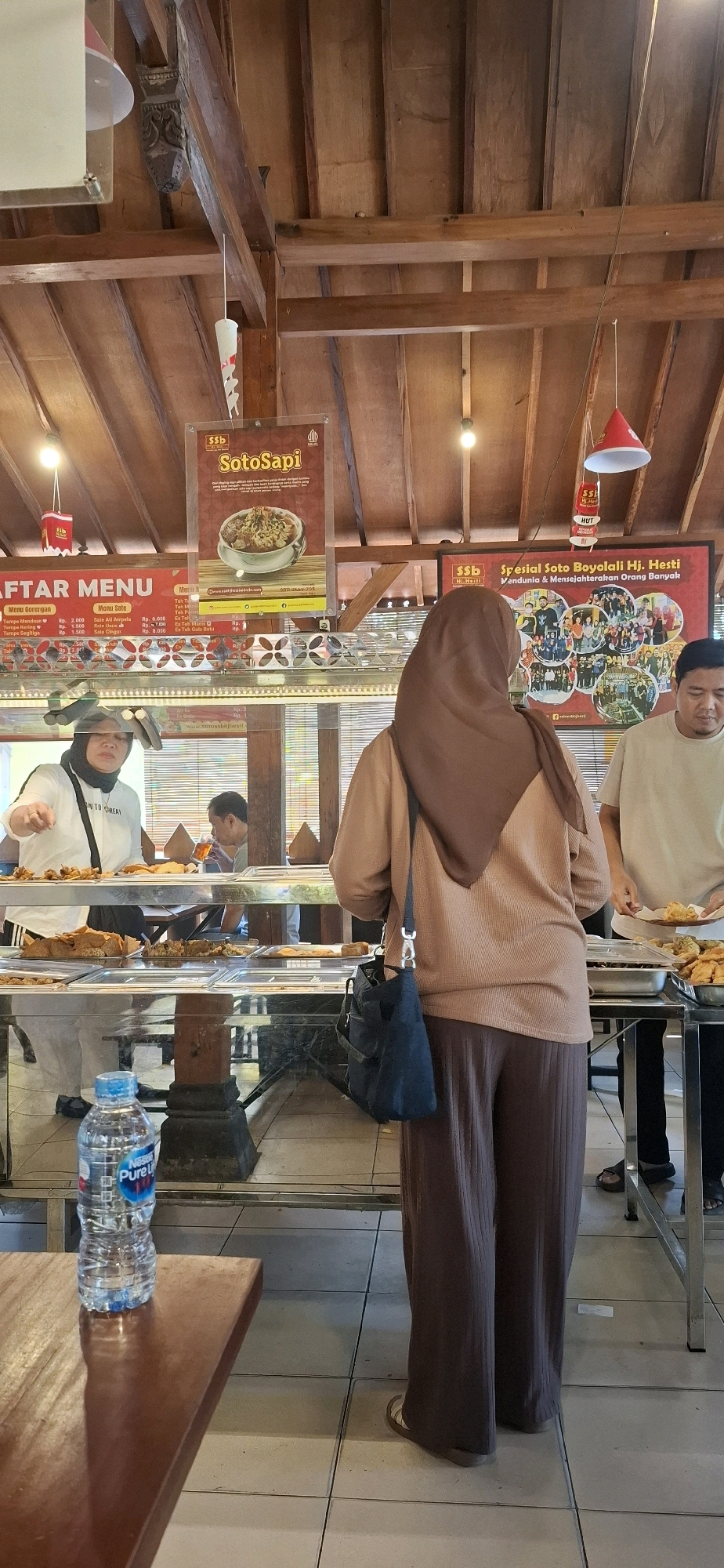 The image captures a scene inside a food establishment in Colomadu, Indonesia, likely a restaurant or a food stall, characterized by its rustic wooden ceiling and visible wooden beams. In the foreground, a clear plastic bottle of Nestle Pure Life water is prominently placed on a dark wooden table.
The midground shows a buffet-style food display behind a glass counter. Several dishes are visible, including what appears to be fried food items and possibly some vegetable dishes. To the left, a woman wearing a white shirt and a headscarf is seen from the chest up, reaching towards the counter, possibly serving herself or selecting food. Behind her, another person is partially visible, seated and holding a glass.
In the center and right of the image, a woman wearing a brown hijab and a long brown outfit is standing with her back to the camera, facing the food display. She has a dark blue bag with a silver clasp slung over her shoulder. To her right, a man in a light-colored t-shirt is standing and holding a plate with food.
Above the food display, several posters are mounted on the wooden ceiling. One poster advertises "Soto Sapi" with an image of a bowl of soup. Another larger poster displays "Spesial Soto Boyolali Hj. Hesti" and a collage of photographs, suggesting a local specialty and possibly the proprietor. The overall environment suggests a casual dining setting.