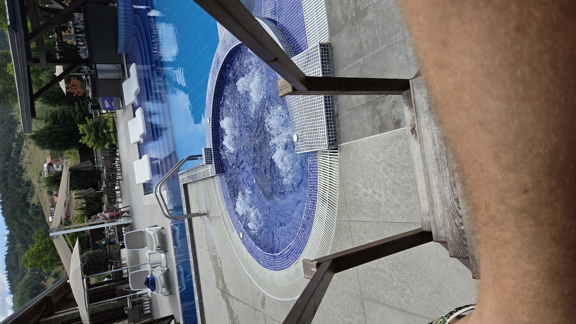 An outdoor view shows a swimming pool and jacuzzi area, likely at a resort. In the foreground, a wooden staircase with a dark brown railing leads down towards the pool area. A person's arm and leg are visible at the very bottom of the frame, suggesting the photo is taken from the perspective of someone using the stairs.
The jacuzzi is circular with blue and white tiled borders, and bubbles are visible in the water, indicating it is active. Adjacent to the jacuzzi is a larger, rectangular swimming pool with blue tiles. Several white, modern lounge chairs are submerged in the shallow end of the pool.
In the middle ground, there is a paved area with tables and chairs set up, some covered by large, light-colored umbrellas. A bar area with bottles displayed is visible to the right, under a wooden canopy. In the background, a lush green, tree-covered hillside with scattered houses forms the backdrop, under a partly cloudy sky. Two women are standing near the tables and umbrellas in the middle ground. The overall scene depicts a leisure facility in a natural, hilly setting.