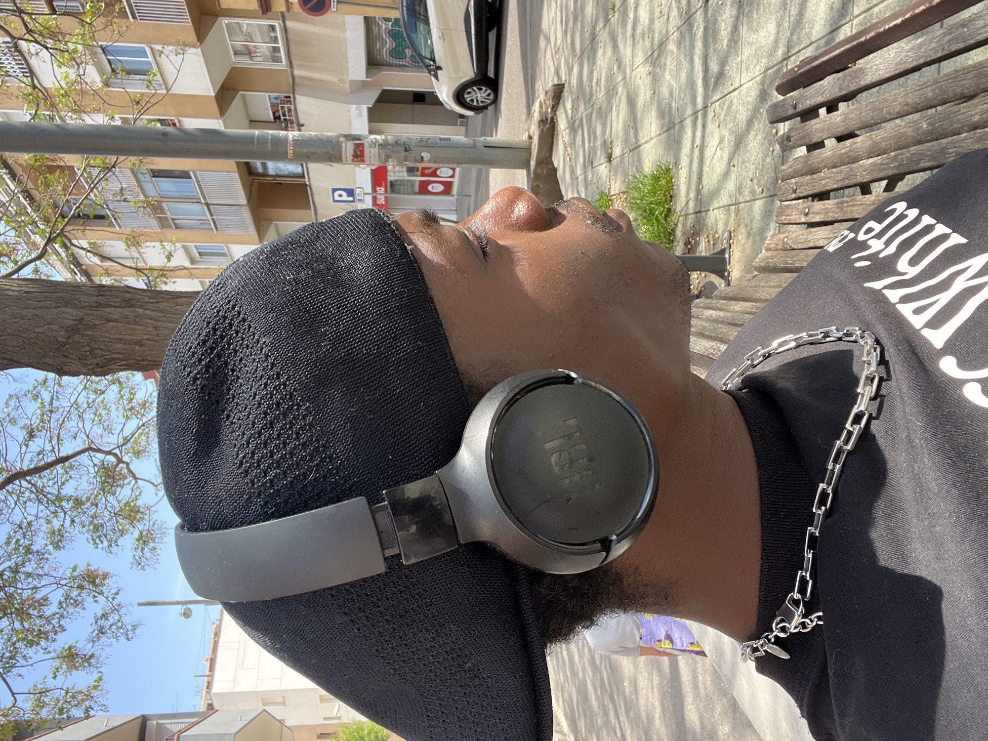 The image depicts a person of color, seen in profile, lying on a wooden park bench and looking upwards with their eyes closed or partially closed. They are wearing a black, textured cap and black JBL over-ear headphones. A silver chain necklace is visible around their neck, and they are dressed in a black t-shirt with white, stylized text, part of which reads "WHITE."
The scene is set in an urban environment during a bright, sunny day, indicated by clear shadows. The background features residential buildings with multiple windows and balconies, characteristic of Barcelona, Spain. A tree trunk and green foliage are visible, along with a lamppost and a white car parked on the street. Various signs are present, including a blue square "P" sign for parking and a red sign with "Suns" or "Suñs" visible. The individual appears to be relaxing or resting, possibly listening to music.