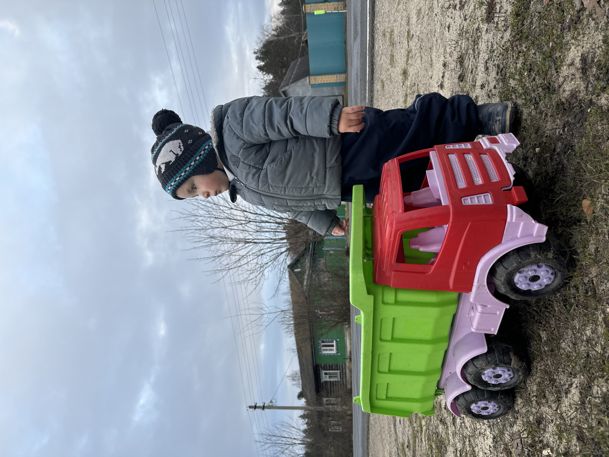 A young child wearing a gray winter coat and a black and blue knit hat with a white animal design is sitting on a large, colorful plastic toy truck. The truck is red, green, and pink. The child is looking down at the truck, possibly playing with it. The location appears to be outdoors near a rural or suburban area, indicated by a gravelly or sandy ground and the presence of trees and a small house in the background. The sky is overcast. The image suggests a cold or cool day. While Красный Маяк in Russia cannot be verified solely from this image, the style of the house and the overall setting could potentially align with rural areas in Russia.