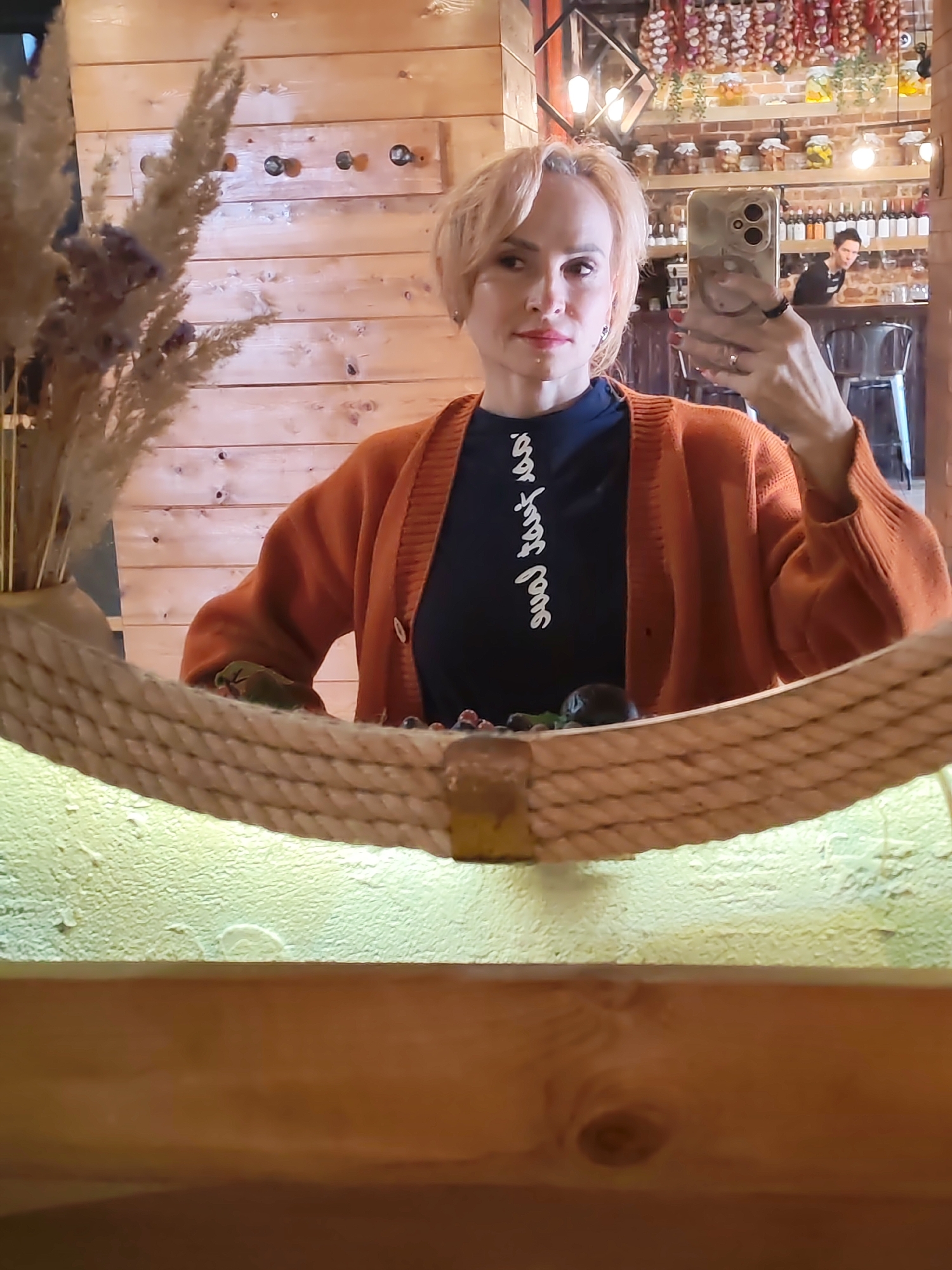 A woman with short blonde hair, wearing an orange cardigan over a dark shirt with white cursive writing, takes a selfie in a mirror. The mirror is framed with thick rope and the background appears to be a rustic wooden wall. In the reflection, shelves with jars and bottles are visible, along with another person in the background. The scene is likely an indoor establishment with a decorative, possibly food-related theme.