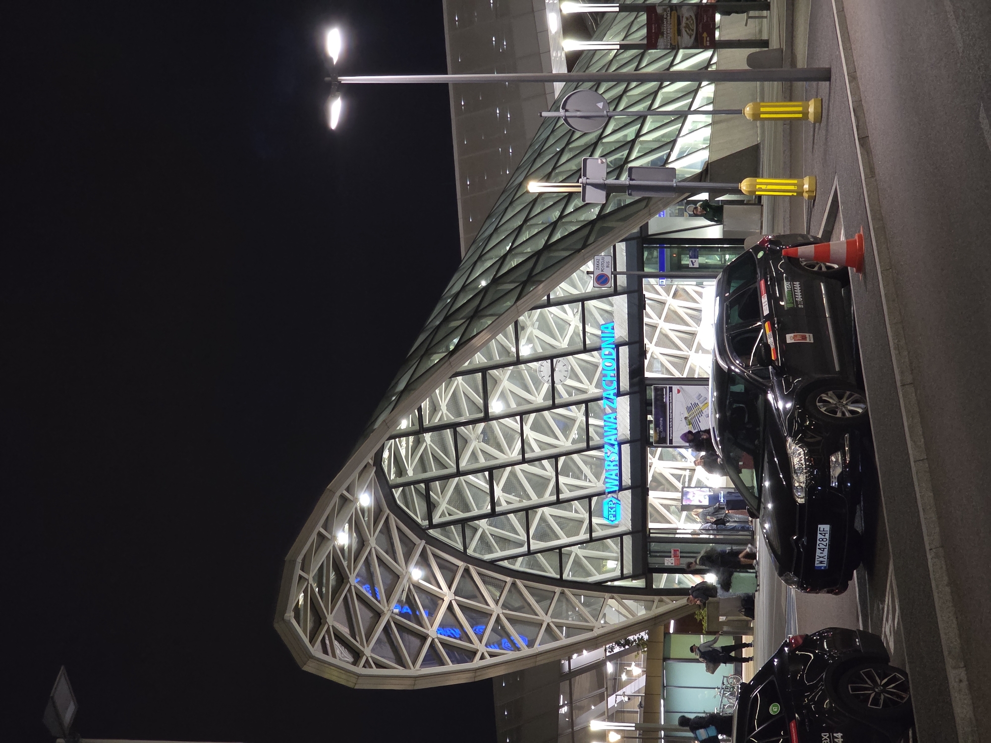 This is a night-time photograph of the PKP Warszawa Zachodnia railway station in Warsaw, Poland. The station features a modern, curved architectural design with a glass and steel facade illuminated by interior lighting and exterior streetlights. Two black cars, possibly taxis, are parked in the foreground on a paved area. One car, a Citroen, has the license plate WX 4284F. The other car, on the left, is partially visible and has "TAXI" branding. People are seen walking in and around the station entrance. The scene is illuminated by artificial lights, creating a somewhat dramatic atmosphere. The sky is dark and clear.