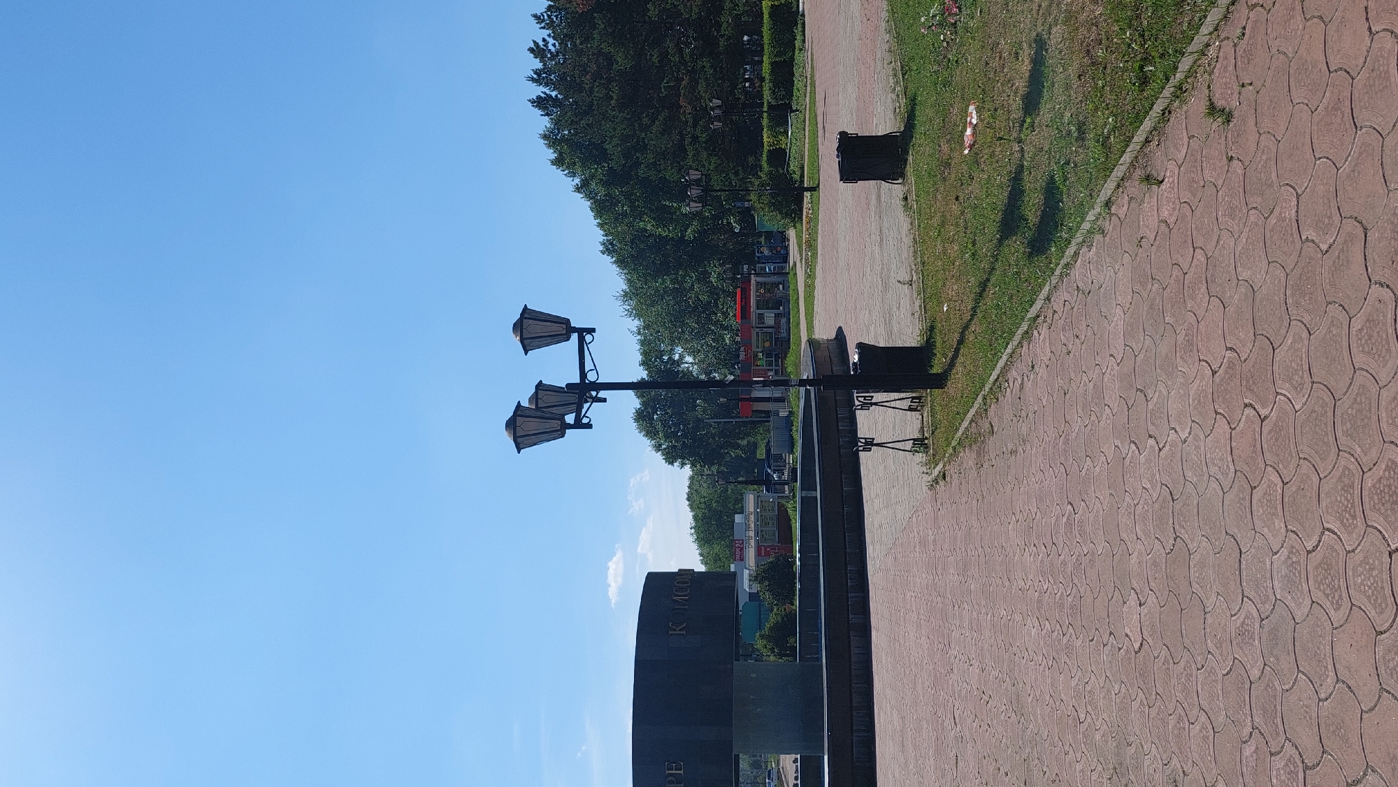 The image depicts an outdoor public space in Komsomolsk-on-Amur, Russia, on a clear day with a bright blue sky. In the foreground, a red, patterned brick walkway leads away from the viewer. To the left of the walkway is a large, dark, cylindrical monument with engraved text, some of which reads "PE" and "КОМСОМ". This monument appears to be part of a fountain structure, though the water is not visible. A tall, black lamppost with three lights stands in the middle of the walkway, and a black trash bin is located to its right. Beyond the walkway and monument, a grassy area is visible, with more trees and some low bushes in the background. Further back, several small retail structures, possibly kiosks or small shops, are visible, with awnings and signage. The overall scene suggests a park or public square area. There are no people visible in the image.