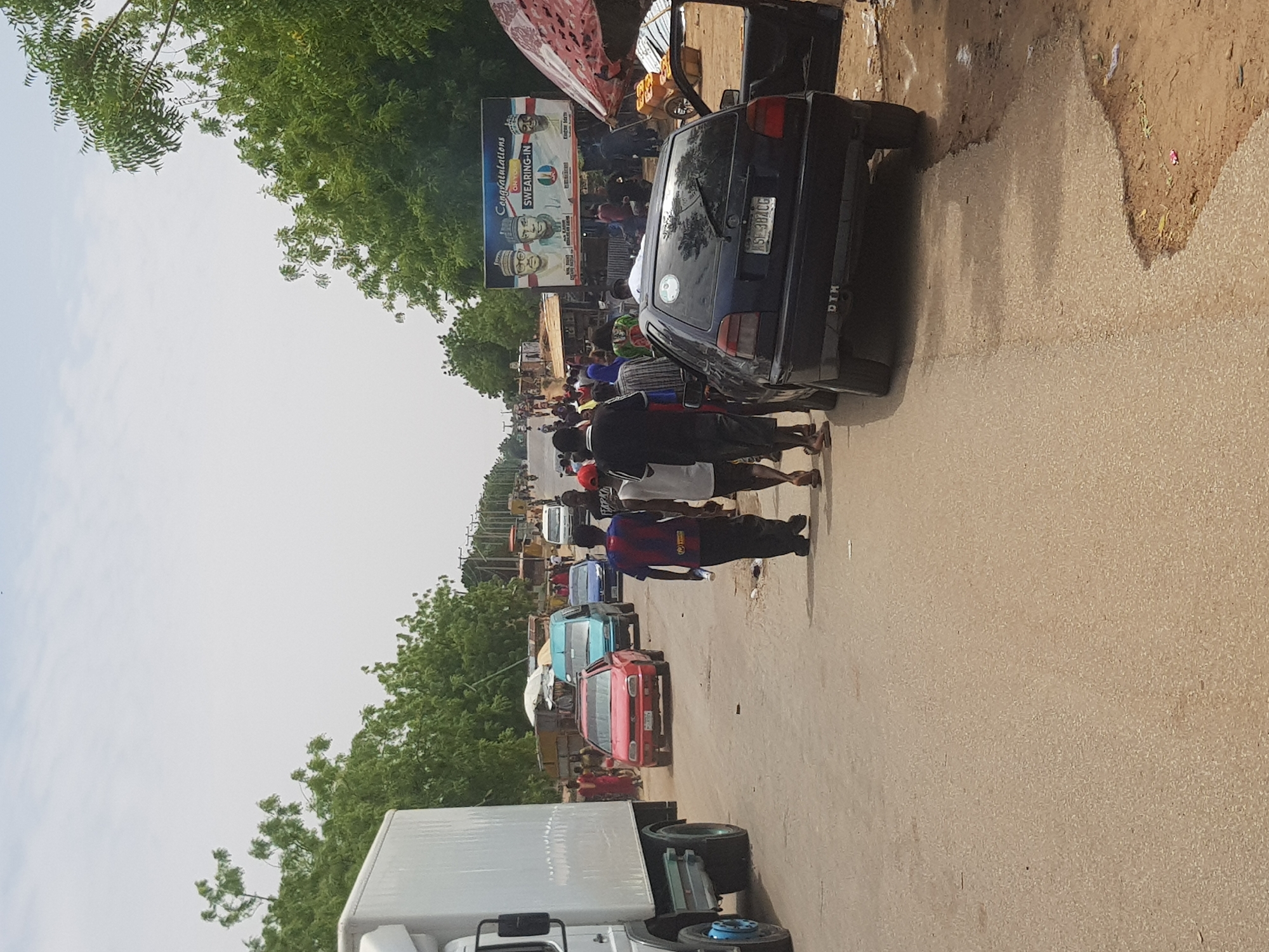 The image shows a street scene in Dabawa, Nigeria. The road is lined with green trees and several cars, including a blue sedan, a red hatchback, and a white truck. Several people are walking on the road. A billboard displaying photos of political figures is visible in the background.