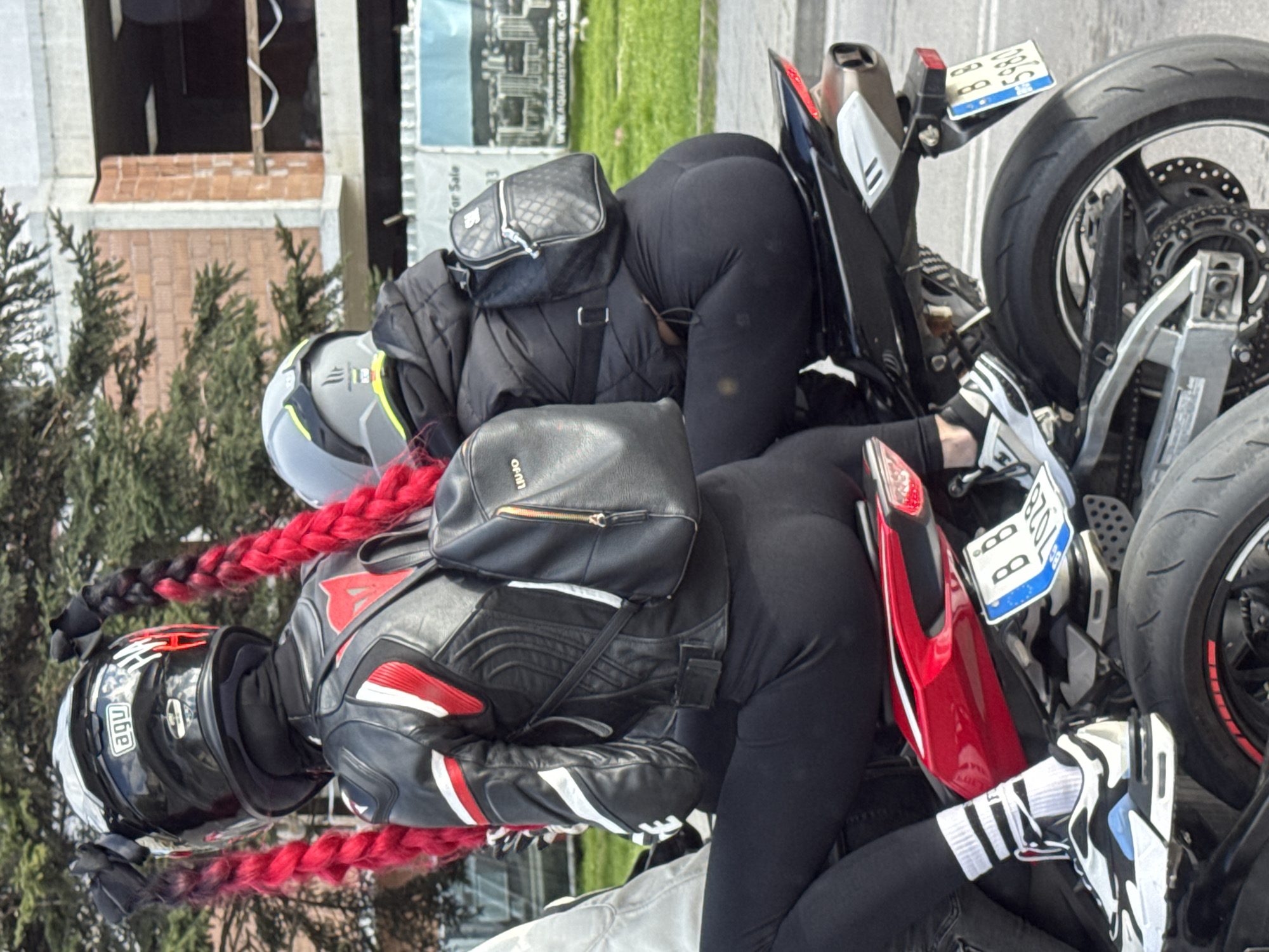 Two individuals wearing motorcycle gear, including helmets, jackets, and black leggings, are positioned next to motorcycles. One individual has red braids. The motorcycles have license plates, one of which is visible and appears to read "BB 7028". The location is indicated as Varna, Bulgaria. In the background, there are trees, a building under construction, and a real estate sign that advertises properties for sale. The entire image is rotated 90 degrees clockwise.