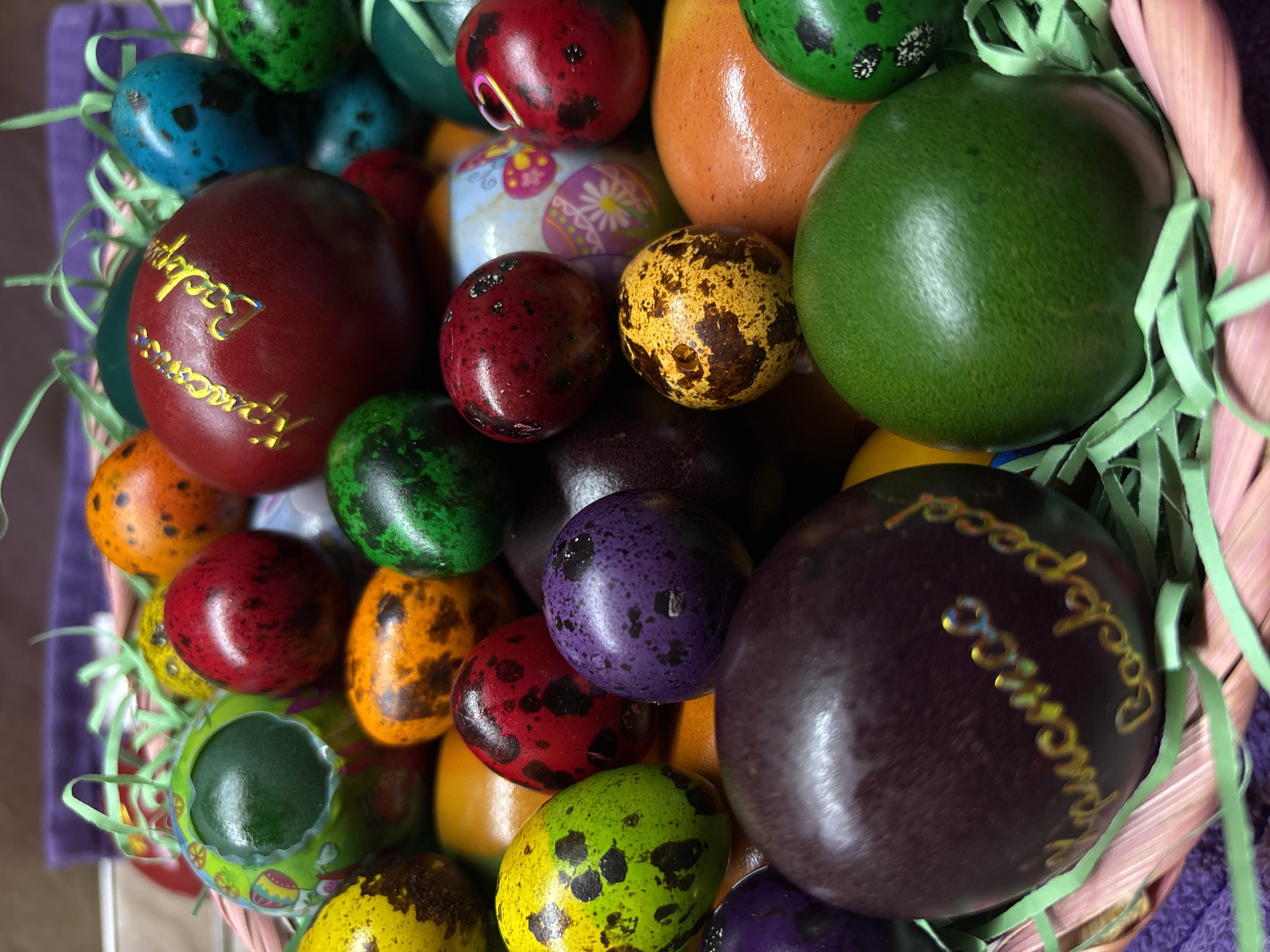 A close-up, high-angle shot depicts a basket filled with Easter eggs of varying sizes and colors. The eggs are predominantly solid colors with some featuring speckled patterns. A few eggs bear inscriptions in gold lettering. The basket is made of a light pink material and is partially filled with green shredded paper. A purple towel is visible in the background. The location is not discernible from the image. No people are visible.