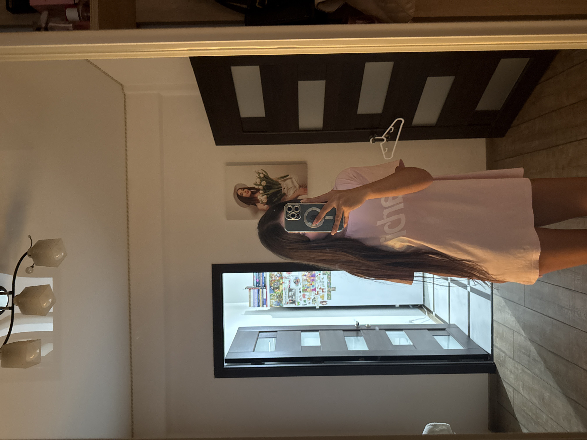 In a mirrored reflection of an indoor setting, a person is visible, holding a smartphone to take a selfie. The person has long, dark hair and is wearing a light pink t-shirt with text that appears to be in a white font. The background of the reflection shows a modern interior with a dark-colored door that has rectangular glass panes. To the left of the door, a framed picture of a woman holding a bouquet of white flowers is displayed on the wall. Further to the left, a lamp with multiple frosted glass shades is mounted on the wall. The floor in the reflection appears to be made of wooden planks. In the foreground, a black-framed doorway with glass panes is visible, leading into another part of the room. The overall impression is that of a well-lit, contemporary living space.