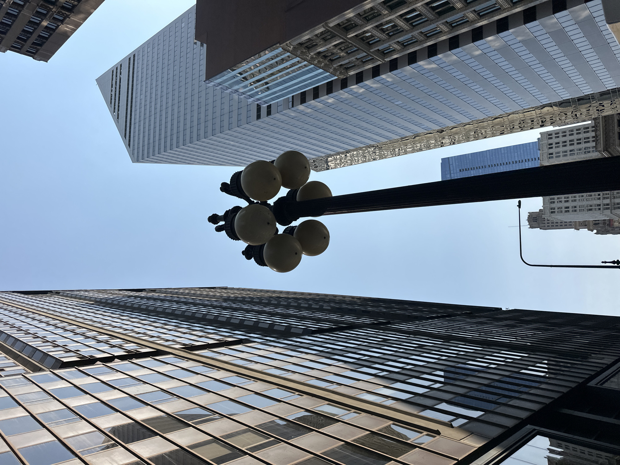 This is a low-angle, exterior, full shot of an urban canyon in Chicago, Illinois. The image is dominated by the reflections of skyscrapers on a clear, sunny day. The sky is a pale blue. In the foreground on the left, a dark, glass-clad building rises high. To the right of this building, a tall, slender lamppost with four globe lights is visible. Further to the right, a sharp, modern skyscraper with a triangular peak, clad in reflective glass, stands prominently. Next to it, another building with a more traditional facade, featuring ornate architectural details and numerous windows, is partially visible. In the background, several other skyscrapers of varying heights and architectural styles can be seen. The overall impression is one of a dense, modern cityscape with a clear sky.