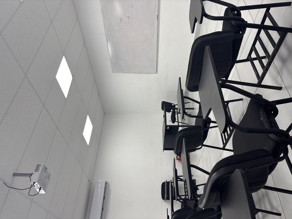 An empty classroom is depicted in Piedras Negras, Mexico. The room features white walls, a light-colored tiled floor, and a suspended ceiling with active square LED panel lights. A white digital projector is mounted to the ceiling near the front left, and a split-system air conditioning unit is installed on the left wall. A large, rectangular whiteboard with faint, illegible markings is mounted on the right wall. The room contains multiple black individual student desks with integrated chairs, arranged in rows. At the front, a black rectangular teacher's desk is positioned with a matching black office-style chair behind it. No individuals are present in the scene, and no actions or interactions are depicted.