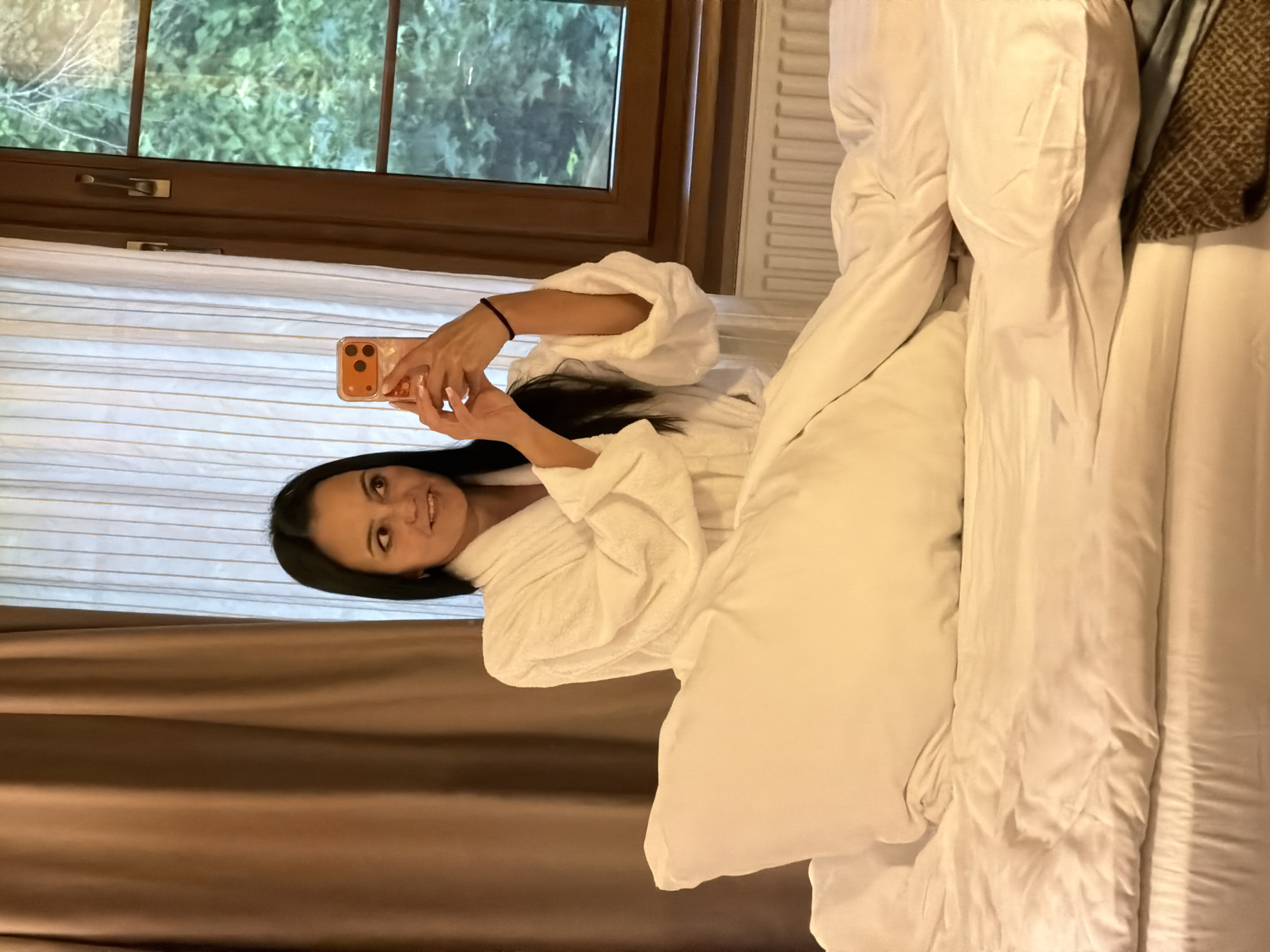 An indoor full-shot photograph depicts a woman in what appears to be a hotel room in Svetlogorsk, Russia. The woman, with dark hair, dark eyes, and a light complexion, is seated on a bed covered with white linens and a white duvet, with a large white pillow positioned in front of her. She is wearing a white terrycloth bathrobe and a dark bracelet on her right wrist. She is holding an orange-colored smartphone with a patterned clear case in both hands, angled towards herself as if taking a selfie. Her expression is a slight smile. To the left of the woman, brown curtains are partially drawn. To her right, a window with a dark brown wooden frame reveals green foliage outside. Below the window, a white radiator is visible. The bed sheets and duvet appear slightly rumpled.