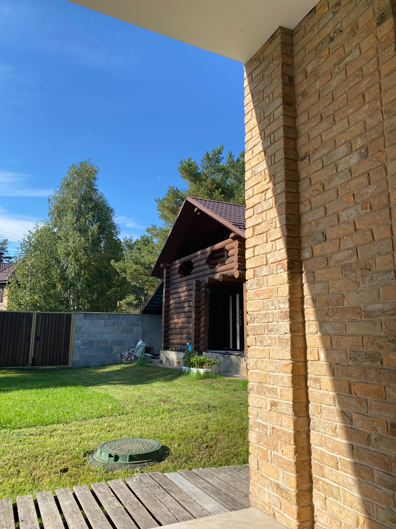 This is a ground-level, eye-level, outdoor shot of a backyard in Perm, Russia. The foreground is dominated by a tan brick pillar, casting a strong shadow on the right side of the image. Beyond the pillar, a wooden walkway leads to a grassy lawn. In the middle ground, a dark wooden fence with vertical planks is visible, bordering a concrete block wall. To the right of the fence is a rustic log cabin with a dark brown roof. Lush green trees, including a tall birch, populate the background, with a clear blue sky overhead. The scene is brightly lit by sunlight, suggesting it is daytime.