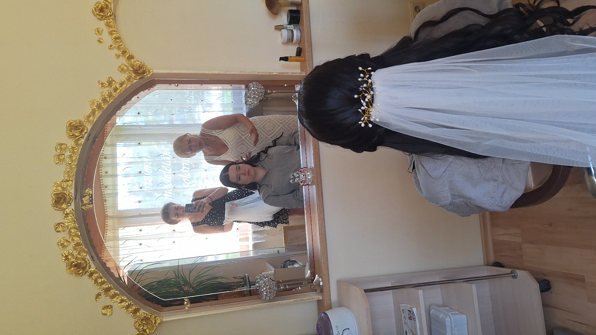 The image depicts an indoor scene, likely a salon or dressing room, with a large, ornate mirror dominating the upper portion. Reflected in the mirror are three individuals and a window with sheer curtains. In the foreground, a person with long, dark hair is seated facing away from the camera, wearing a light-colored jacket and a white veil with a pearl and gold hairpiece. To the left of this person, a shelf unit with various items is visible.
In the reflection, a young woman with dark hair is seated, looking down and holding a mobile phone. Beside her stands another woman in a black and white patterned dress, holding a white garment. To the right of them, an older woman in a white lace dress gestures with her hands. The background reflection shows a window with patterned curtains, and various decorative items are placed on a shelf behind the mirror, including crystal candle holders and bottles of products. The overall ambiance suggests preparation for an event, possibly a wedding. The scene is identified as being located in the city of Ogre, Latvia.