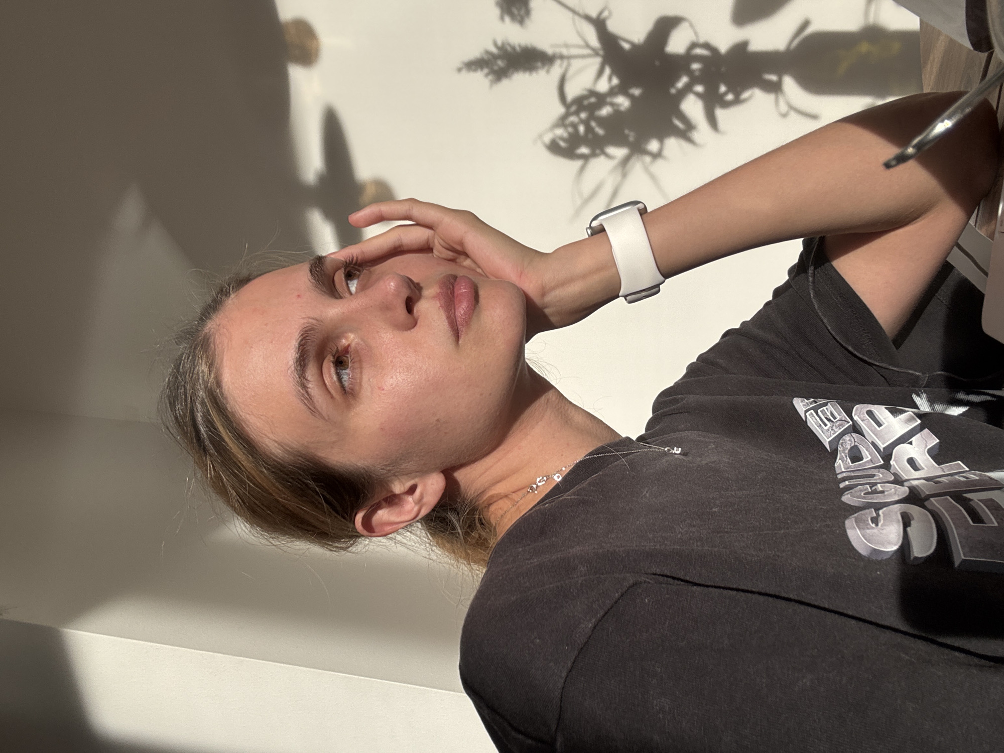 A woman is posing indoors. She has light brown hair and pale skin. She is wearing a dark grey t-shirt. She is wearing a white smartwatch on her left wrist and a delicate silver necklace. She is touching her face with her left hand. There is a vase containing a plant casting a shadow on the white wall behind her. The location is unspecified.