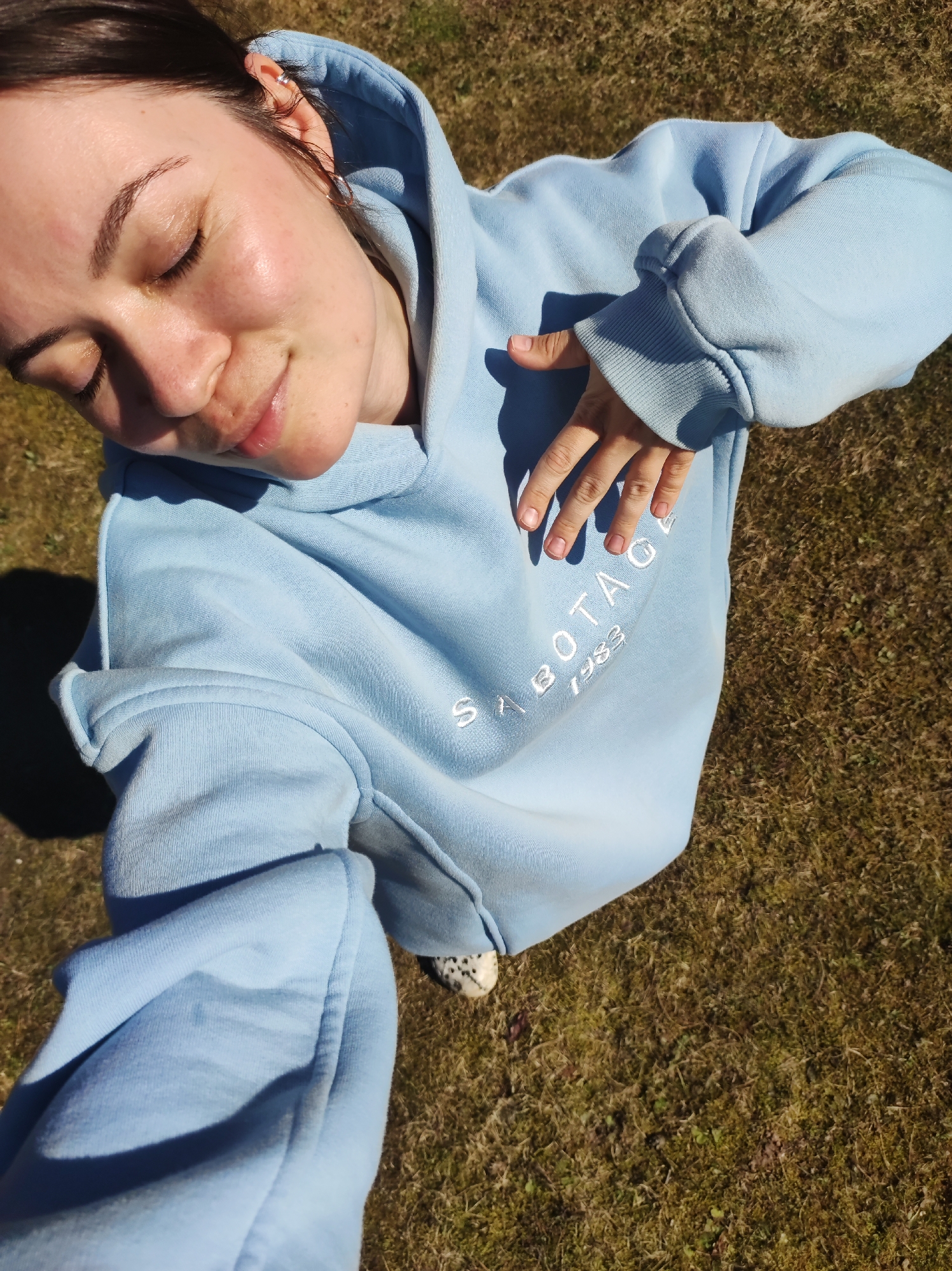 A young woman is taking a selfie outdoors. She is wearing a light blue hoodie with the word "SABOTAGE" and "1983" embroidered on the front. Her eyes are closed and she is smiling slightly. Her hand is resting on the front of her hoodie. The background is brown grass. There is no information in the image to indicate a location in Фоки, Russia.