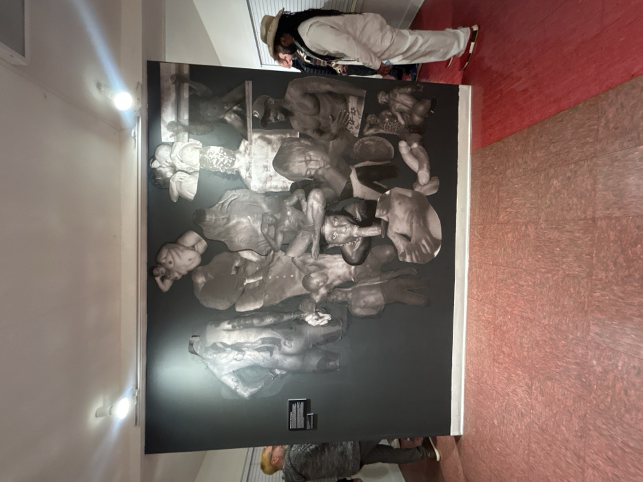 A large, dark wall features a collage-style artwork. It consists of black and white images of human figures and body parts overlapping each other. Two individuals stand near the wall, apparently viewing the piece. The person in the foreground has a light complexion and short hair. Above, a person wearing a light-colored suit and a straw hat examines the artwork closely. The room has a light-colored wall on the left and a red and beige patterned floor.