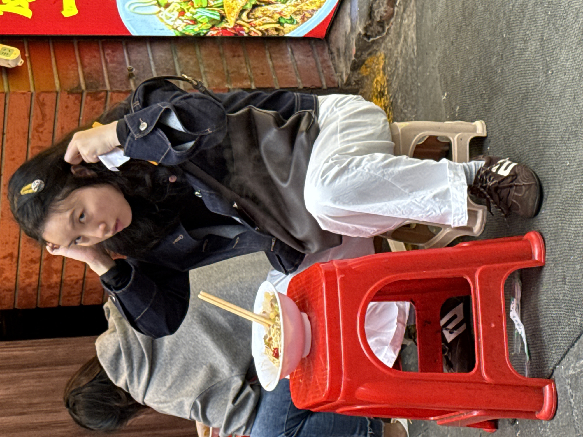 An outdoor scene in Yuzhong District, China, depicts two individuals in a casual dining setting. In the foreground, a young East Asian female is seated on a light beige plastic stool, facing slightly towards the viewer. She wears a black top, a dark denim jacket with rolled-up sleeves, white loose-fitting pants, and dark brown lace-up sneakers with white markings. Her dark hair is adorned with a golden hair clip, and her right hand is raised near her temple, holding a small white tissue or napkin. A dark brown shoulder bag is slung across her body. Directly in front of her, on a bright red plastic stool, rests a white bowl containing noodles, vegetables, and other ingredients, with two wooden chopsticks inserted upright. To her left and slightly behind, another individual with dark hair is visible from the back, wearing a gray hooded sweatshirt and blue jeans, seated and facing away from the viewer. The background features a reddish-brown brick wall on the right, adorned with a tall red sign displaying yellow characters and an image of food. A small, light-colored sensor is mounted on the brick wall above the sign. To the left, a dark brown curtain or wooden panel is visible. The ground consists of a textured dark surface, with a piece of crumpled white paper visible near the base of the red stool.