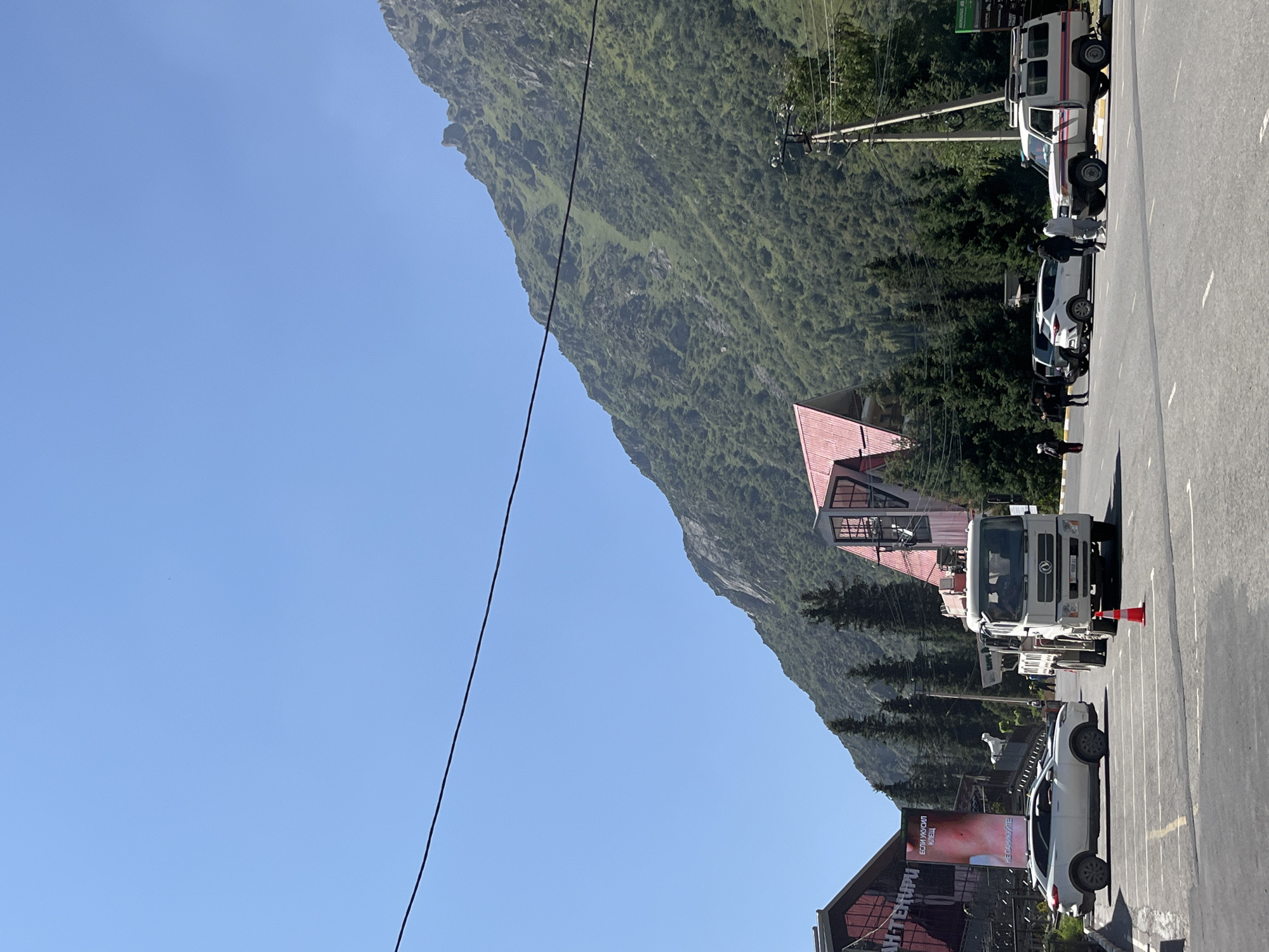 The image depicts a street scene in Kashka Suu, Kyrgyzstan, with a prominent mountainous backdrop. In the foreground, a paved road with white lane markings is visible. Several vehicles are present: a white SUV parked on the left, a white truck with a crane positioned centrally, and a white off-road vehicle parked on the right. A traffic cone is placed in front of the truck. A group of individuals are walking on the road and sidewalk. Buildings with red roofs are situated to the left and behind the vehicles, with signage visible on one of them. Evergreen trees line the area between the road and the mountains, which are covered in lush green vegetation. The sky is clear and blue.