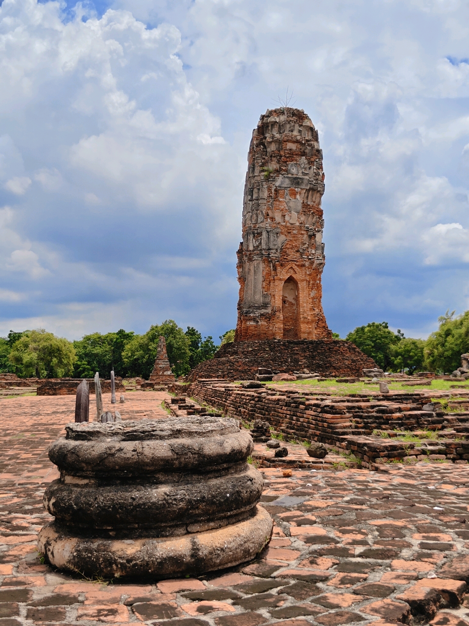 This image depicts a scene at Wat Phra Si Sanphet, located in the Phra Nakhon Si Ayutthaya City Municipality, Thailand. The foreground is dominated by the weathered stone base of what appears to be a fallen pillar, resting on a paved courtyard of red bricks. In the mid-ground, the ruins of ancient brick structures are visible, including walls and a central, tall, cylindrical stupa that is partially collapsed and covered in moss and vegetation. The stupa exhibits typical Ayutthaya architectural features with decorative niches. The background consists of lush green trees and a sky filled with dramatic white and gray clouds, suggesting either an approaching storm or recent rain. There are no people visible in the image. The overall impression is one of historical decay and the enduring presence of a significant ancient site.