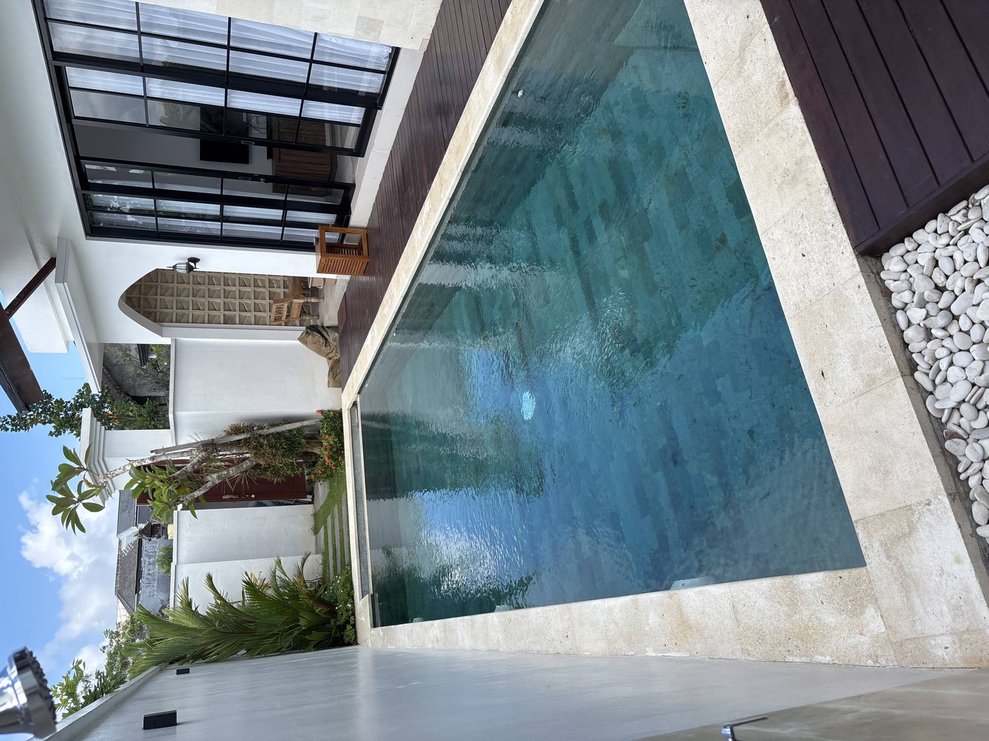 The image depicts an outdoor swimming pool situated in Ungasan, Indonesia, bordered by a white stone edge. The pool's water has a blue-green hue. One side of the pool is flanked by a wooden deck and a row of white stones. Adjacent to the deck is a building with black-framed windows and doors. An outdoor seating area with a woven design can be seen through an arched opening. A tropical landscape with palm trees and other greenery is visible. The sky is blue with scattered clouds. The image is taken from an elevated perspective.