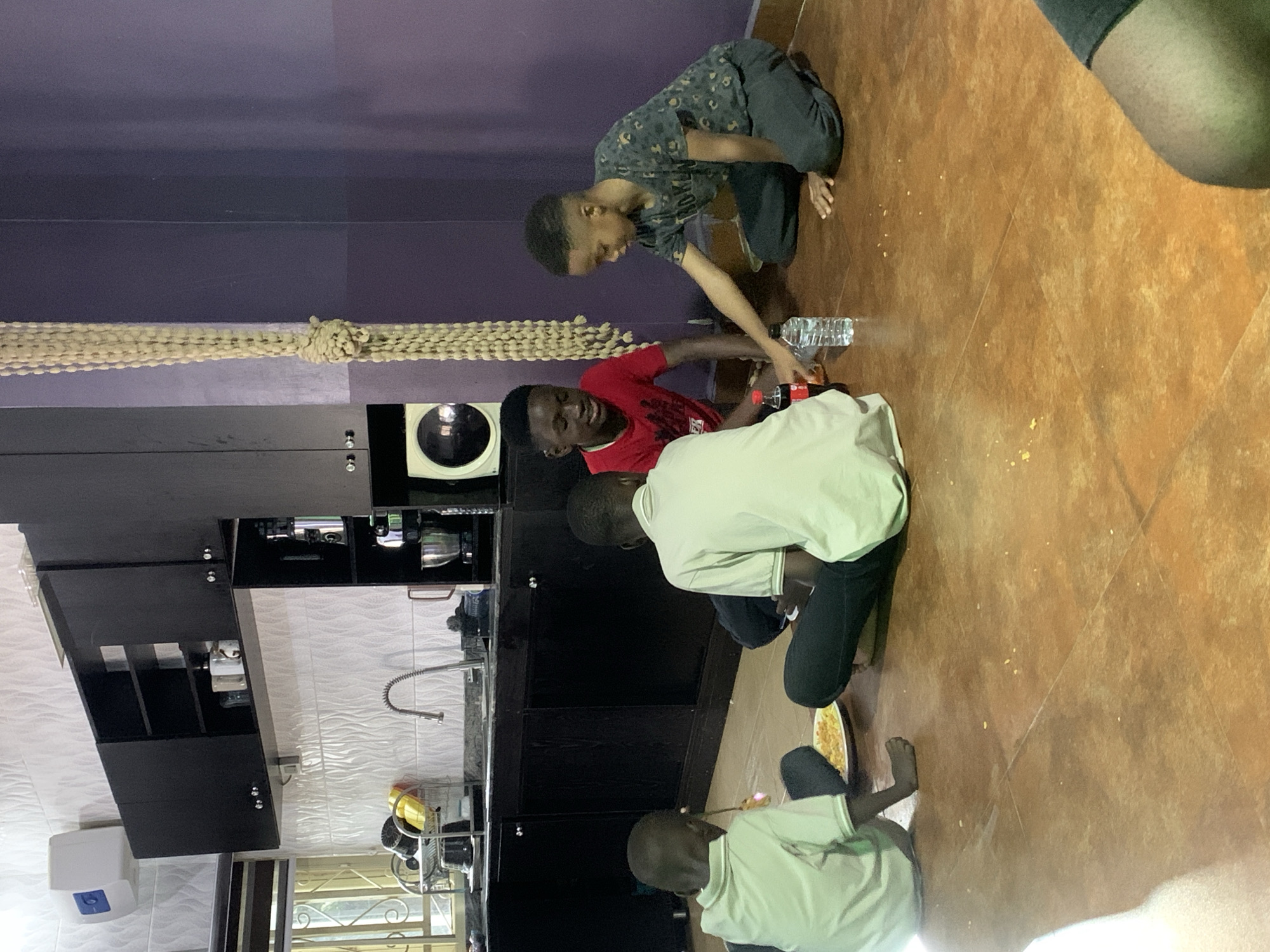 In an indoor setting, identified as a residential space within the city of Jos, Nigeria, four young boys and a partial view of a fifth person are present. The primary scene features four boys seated on a brown tiled floor, indicative of a shared meal or gathering. The background consists of a kitchen area on the left and a plain purple wall on the right. The kitchen features dark, possibly black, upper and lower cabinets with silver handles. A white textured tiled backsplash extends along the wall above the counter. A silver gooseneck faucet is visible above a sink. To the left of the sink, a silver dish-drying rack holds various items, including yellow and black utensils or containers. Above the upper cabinets on the far left wall, a white rectangular appliance, possibly a water heater or storage unit, is mounted. Below the upper cabinets on the right side of the kitchen, a small white appliance with a circular dark front, resembling a microwave or mini-oven, is visible, next to a silver mixer bowl. Centered on the right side of the image, separating the kitchen from the purple wall, a long, light beige or off-white macrame or woven rope hangs vertically, secured by a knot at mid-height. Regarding the individuals: * On the far left, a boy in a light green polo shirt and dark trousers sits with his back partially to the viewer, facing left. A white plate containing yellow food, possibly rice or pasta, is on the floor in front of him. He holds a small piece of food in his right hand near his mouth. * To his right, a second boy in a light green polo shirt and dark trousers sits facing away from the camera, looking towards the right side of the image. * Next to him, a boy in a red t-shirt with a white graphic (partially obscured) sits facing right, smiling. He looks towards the boy reaching for the bottle. * Opposite the red-shirted boy, a fourth boy in a dark patterned (possibly camouflage) t-shirt and dark trousers kneels, facing right. He extends his right arm towards a clear plastic water bottle on the floor. * Between the red-shirted boy and the patterned-shirted boy, a small red plastic Coca-Cola bottle is also on the floor. * Scattered small yellow crumbs are visible on the brown tiled floor around the boys. * In the
