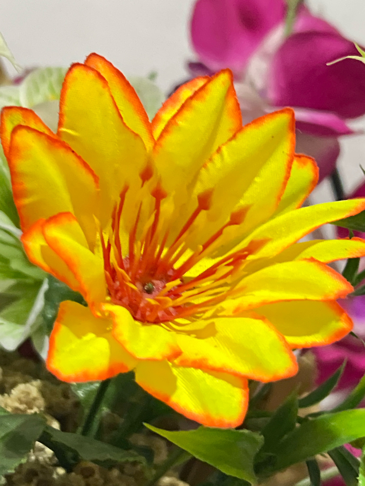 The image displays a close-up of a vibrant yellow and orange flower, likely artificial, with numerous delicate petals. The flower's center features a cluster of thin, red stamens. In the background, out-of-focus elements of other artificial flowers are visible, including a large, pinkish-purple bloom. The surrounding foliage appears to be green and also artificial, with some texture suggesting it's part of a decorative arrangement. The lighting is soft, creating gentle shadows on the petals. The scene suggests an indoor decorative setting, possibly in a hotel, shop, or private residence. The specific location in Denpasar, Indonesia, is not discernible from the visual information provided in the image.