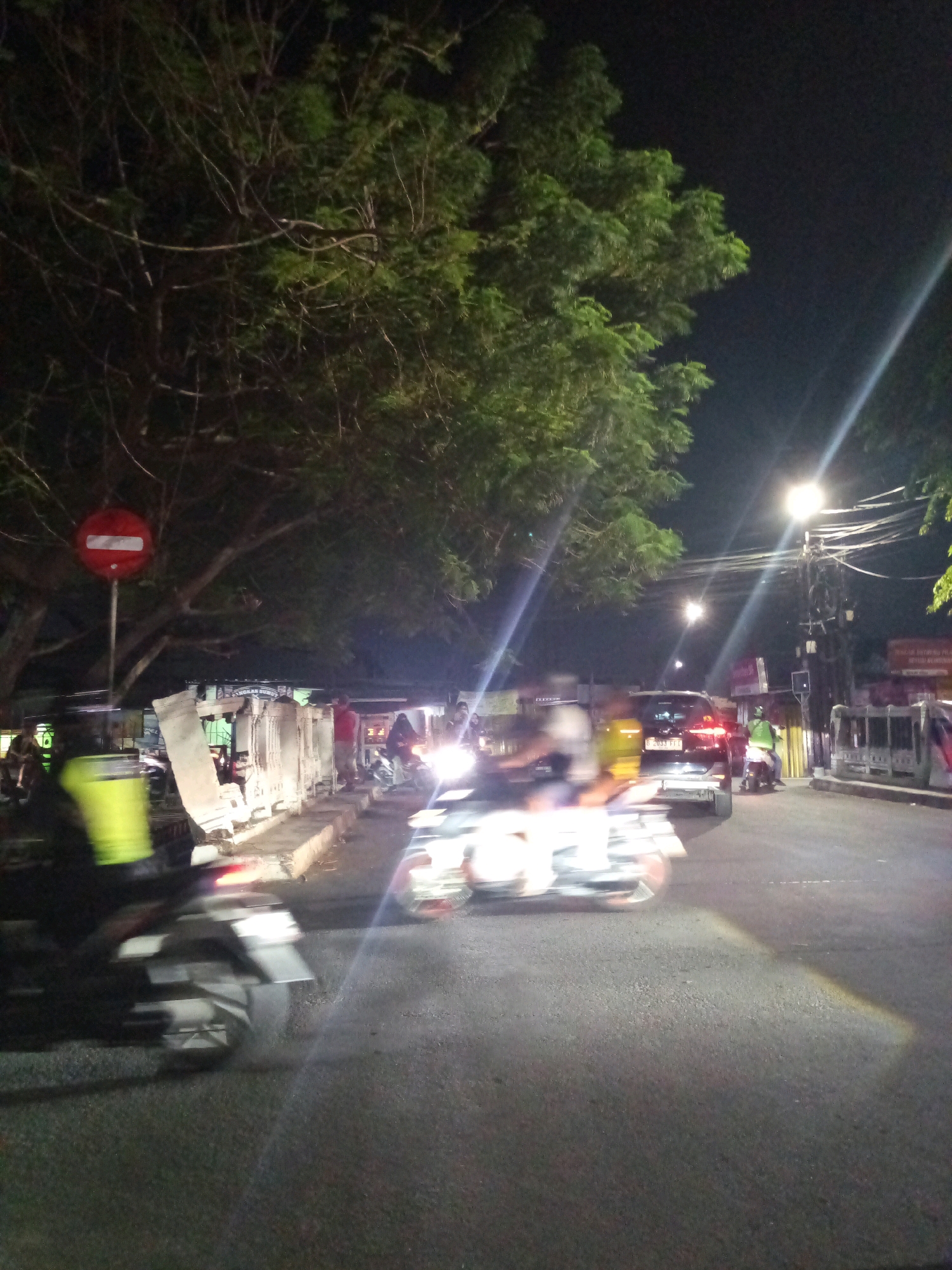 This is a nighttime, slightly blurred photograph taken on a street in Bekasi, Indonesia. A tree with green foliage dominates the upper left portion of the frame, with its branches extending over a sidewalk and road. A red circular sign with a white horizontal bar, indicating a prohibition, is mounted on a pole near the tree.
The scene appears to be a busy street with vehicular traffic. Several motorcycles are visible, with their headlights and taillights creating streaks of light due to motion blur. One motorcycle in the foreground on the left has a rider wearing a bright yellow vest, also blurred. Another motorcycle is seen in the mid-ground on the right, with its rider also obscured by motion.
A dark-colored SUV with license plate B 2033 FIT is stopped or moving slowly in the right lane. Pedestrians are present on the sidewalk, some standing and some walking. Various structures line the sidewalk, including what appear to be shops or stalls, some with lights on. Overhead, streetlights and power lines are visible against the dark night sky. The road surface is asphalt. The overall impression is of a lively urban environment at night.