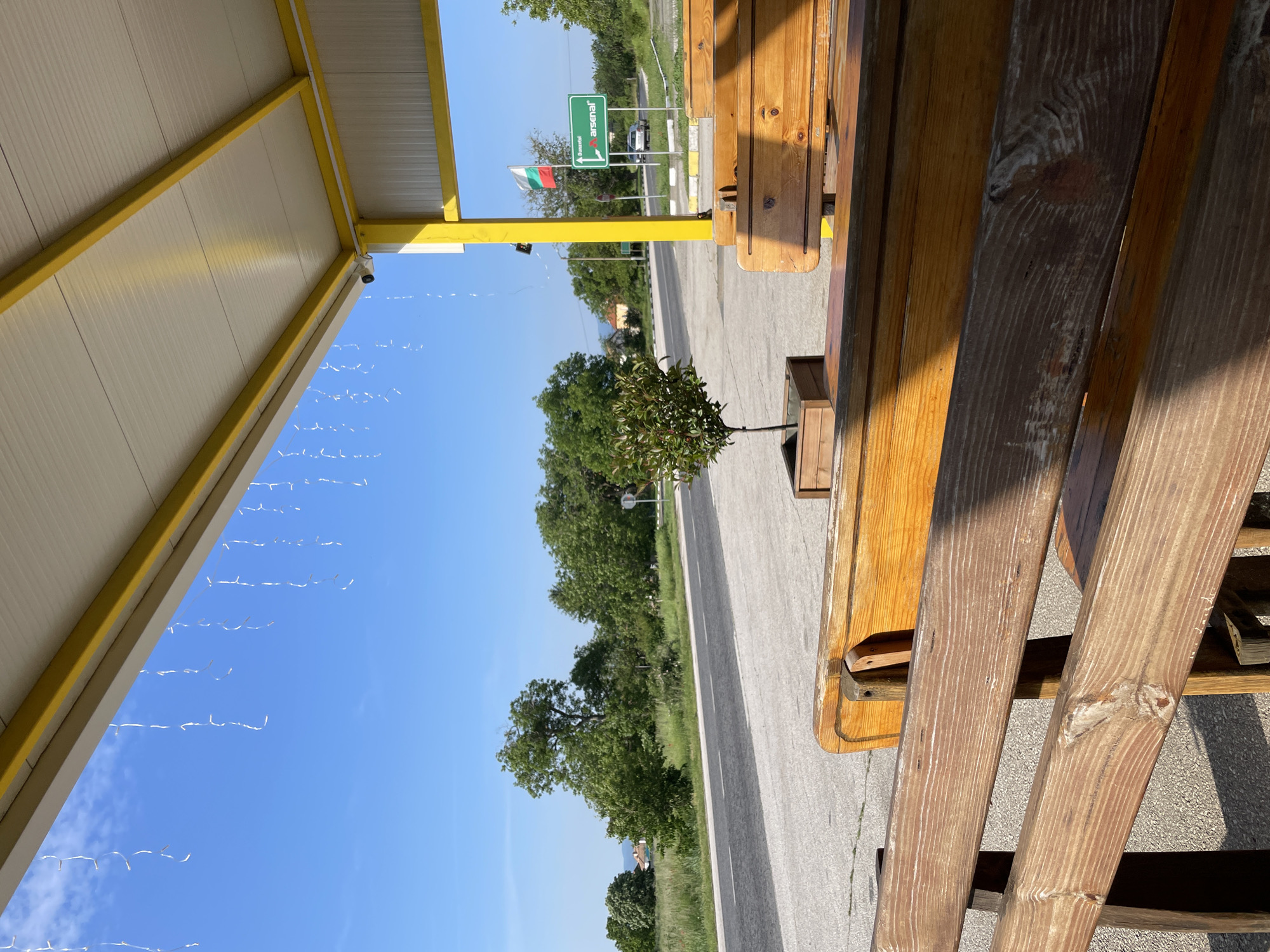 A daytime scene shows a roadside rest area in Sheynovo, Bulgaria. A yellow-trimmed shelter roof covers a picnic table with wooden benches, providing shade. A small tree in a planter sits near the edge of the table. A road stretches into the distance, flanked by green vegetation. A road sign with "Arsenal" in green letters and a Bulgarian flag is visible. A camera is attached to the roof. The sky is clear and blue, with a strand of lights hanging from the roof.