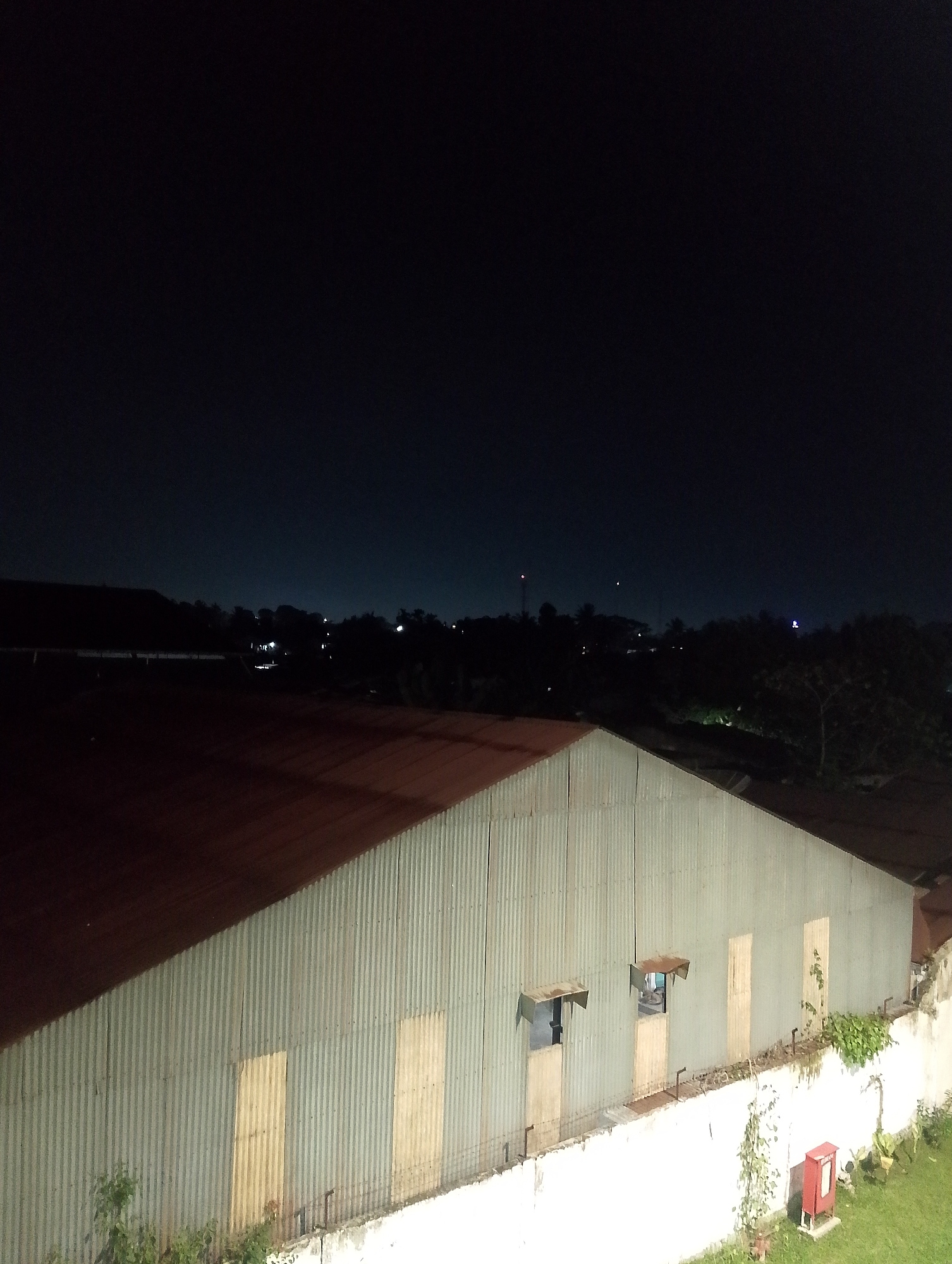 The image shows a corrugated metal warehouse at night in the city of Pematang Siantar, Indonesia. The warehouse has a dark red, sloped roof and a light gray, corrugated metal facade. There are several boarded-up openings on the facade, appearing as light brown rectangles. One opening on the left side has a small, metal awning. Another opening further to the right appears to be a window or doorway with a blue light visible inside. A white concrete wall runs along the front of the warehouse, with some green vegetation growing on it and along the ground in front. A red fire alarm box is mounted on a metal post near the wall. In the background, a dark sky with some distant lights suggests a city skyline. The overall scene is industrial and appears to be taken from an elevated vantage point, possibly a balcony or another building.