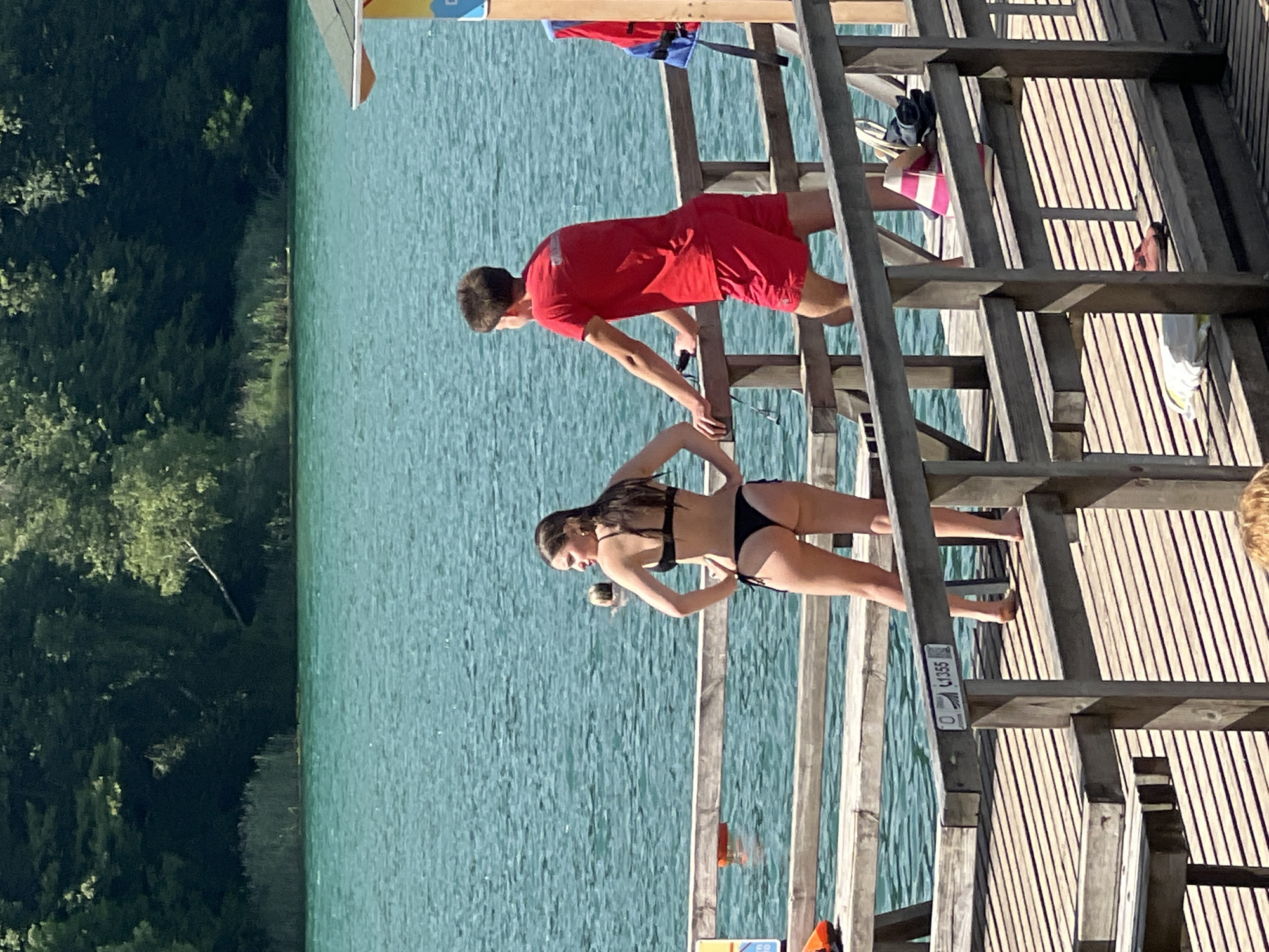 In Vilnius, Lithuania, on a sunny day, a young woman in a black bikini stands on a wooden pier, facing away from the camera with her hands on her hips. Beside her, a young man in a red t-shirt and red shorts leans on the pier railing, looking out at the water. The turquoise water of a lake stretches out before them, bordered by a dense green forest. Two orange buoys are visible in the water. The pier is constructed of weathered wooden planks and railings, with a few items resting on it, including a blue and red life jacket, a pink and white striped object, and a pair of shoes.
