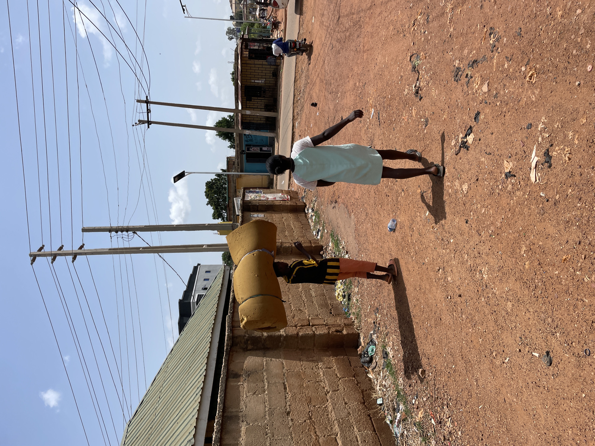 The image depicts a street scene in Jos, Nigeria. In the foreground, a child is carrying a large, yellow mattress roll on their head, secured with rope. They are wearing a dark, horizontally striped shirt and peach-colored shorts. To their right, an adult woman in a light green dress is walking ahead, with her right arm raised slightly. The ground is reddish-brown dirt, littered with debris. A brick wall runs along the left side of the frame, topped with a green corrugated metal roof. Power lines stretch across the sky above. In the background, a motorcycle and other buildings are visible.