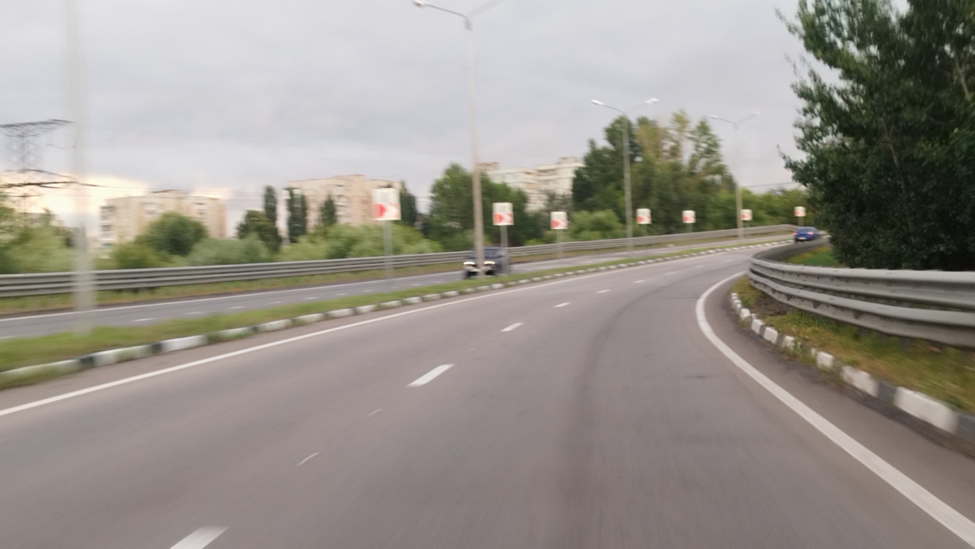The image captures a highway scene in Stary Oskol, Russia, under an overcast sky. The perspective is from a vehicle moving along the road, indicated by motion blur. The highway curves to the right, bordered by a metal guardrail on the outer edge. On the left side of the highway, there are apartment buildings in the distance, some appearing to be several stories high, interspersed with trees and vegetation. Streetlights are visible along the side of the road, as are traffic signs, specifically square signs with red arrows pointing to the right, indicating a curve or turn. Two vehicles are partially visible on the road: a dark-colored car in the middle distance on the left lane, and a blue car further ahead on the right lane. The road surface is asphalt, marked with white lane lines. The overall environment suggests an urban or suburban highway setting on a cloudy day.