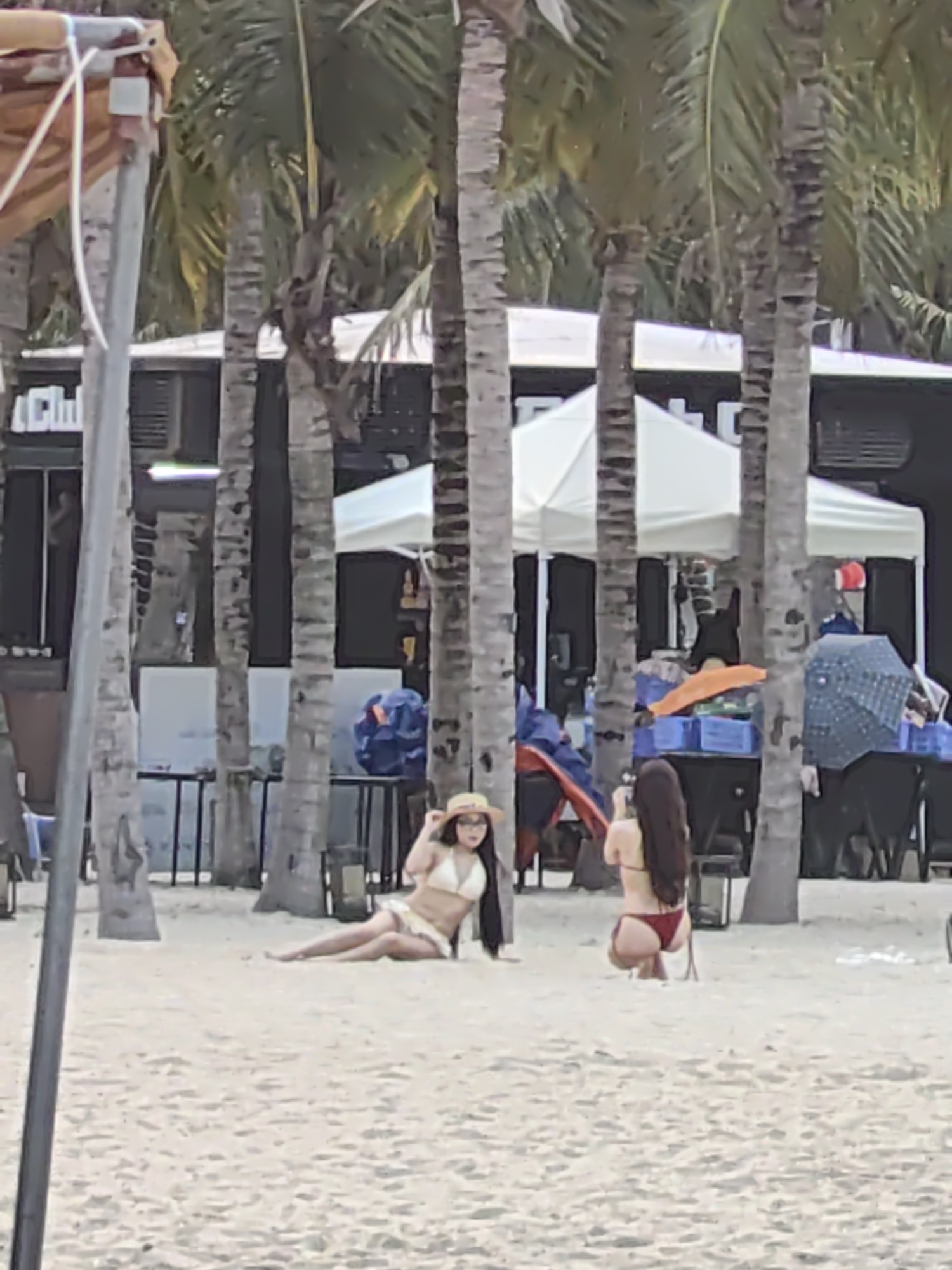 The scene is a beach in Bai Chay Ward, Vietnam. Two women are on the sand. One woman is lying down, wearing a white bikini and a hat. The other woman is kneeling and taking a photograph of the first woman with a smartphone. There are palm trees behind the women. In the background, there are buildings with awnings, tables, chairs, and other people under the awnings. Also, a blue beach bag, an orange towel, and blue crates are present in the background.
