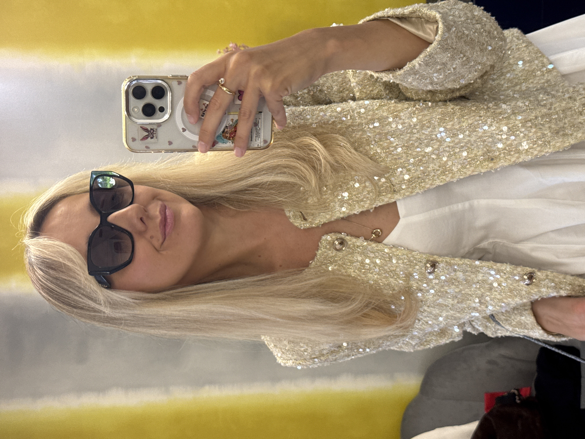 A blonde woman is taking a selfie in a room with a yellow and gray striped wall. She is wearing large black sunglasses, a gold sequined jacket, and a white top. She has a gold necklace with a small pendant. On her right hand, she is wearing a gold ring with a small pearl. She is holding a smartphone which has stickers on its case. To her left, a grey couch is visible.