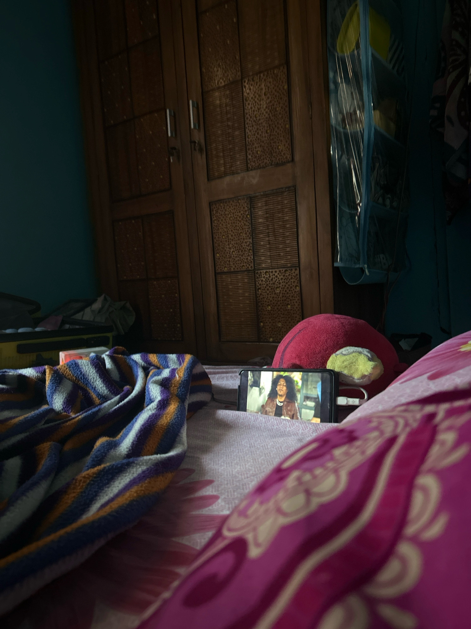 In a bedroom in Cihaurbeuti, Indonesia, a smartphone is placed on a bed, displaying a video of a person with dark, curly hair wearing a brown jacket and a black shirt. The phone is positioned on a pink and white patterned sheet, with a colorful striped blanket draped over the left side of the bed. A red spherical pillow and a yellow and green object are also on the bed behind the phone. In the background, a dark wooden wardrobe with intricate patterns is visible, along with a blue wall and a hanging organizer containing yellow items.