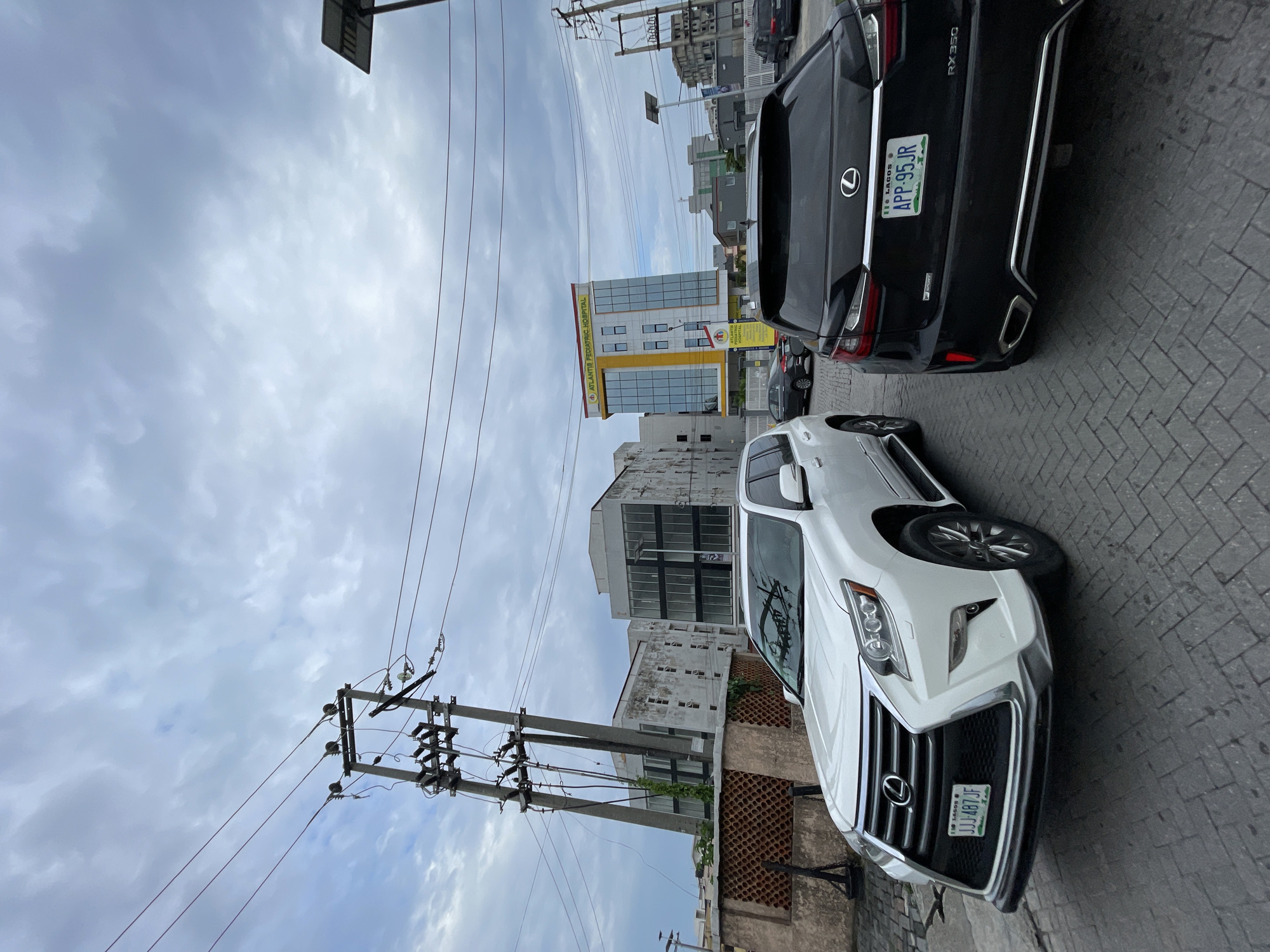 This media file depicts a street scene in Moba, Nigeria, with a focus on two parked vehicles and a building. The primary subjects are a white Lexus SUV in the foreground, angled towards the viewer, and a black Lexus SUV behind it. The white SUV has its license plate, "JJJ-487 JF", clearly visible, which indicates "LAGOS". The black SUV's license plate, "APP-95JR", also indicates "LAGOS" and the model "RX350".
The scene includes architectural elements, notably a modern building with a yellow and glass facade, identified by signage as "ATLANTIS PEDIATRIC HOSPITAL". Several utility poles with numerous wires are prominent in the background and foreground, suggesting an urban or semi-urban environment. The ground is paved with brickwork.
The sky is overcast with heavy clouds, indicating a cloudy or potentially rainy day. The time of day is indeterminate due to the lighting, but the overall atmosphere is neutral. There are no people visible in the frame. The composition is such that the image is oriented vertically, as if taken with a phone held upright. The prominent Lexus vehicles and the hospital building are the main points of interest.