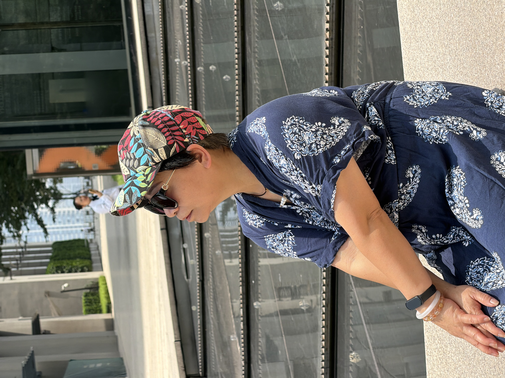 In Yuzhong District, China, a woman wearing a navy blue patterned outfit and a colorful patterned cap with sunglasses is sitting down outside. She is looking up and to her right. She has bracelets on her left wrist, one made of clear beads and another that is black. Behind her, there is a building with reflective windows and a tree. In the distance, another person in a white jacket is visible.