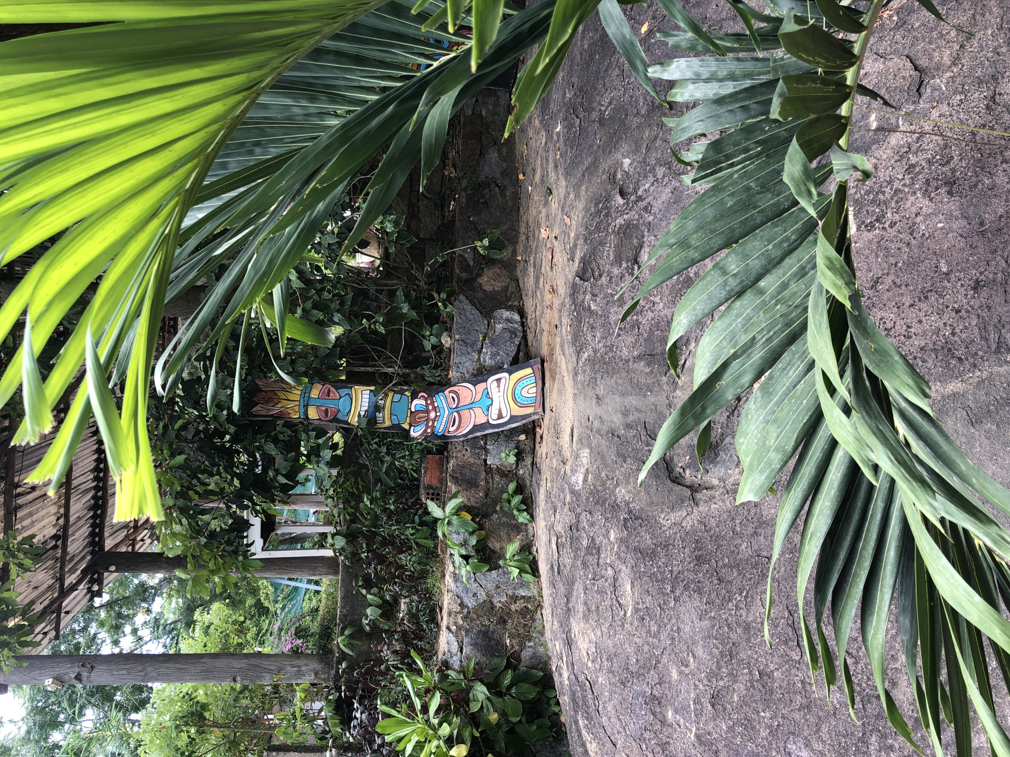 The image depicts a colorful tiki pole leaning against a stone wall, located in a lush garden setting. The tiki pole is decorated with stacked faces, each with distinct expressions and vibrant colors, including a rainbow at the bottom. Behind the tiki pole, there is dense foliage and vegetation, with a partially visible wooden structure overhead, suggesting an outdoor seating area or a gazebo. A large, textured boulder occupies the foreground, with large palm fronds obscuring parts of the view. The overall scene conveys a tropical and somewhat rustic atmosphere, likely an outdoor space in a city like Da Nang, Vietnam, known for its beaches and tropical climate. There are no people or animals present in the image.
