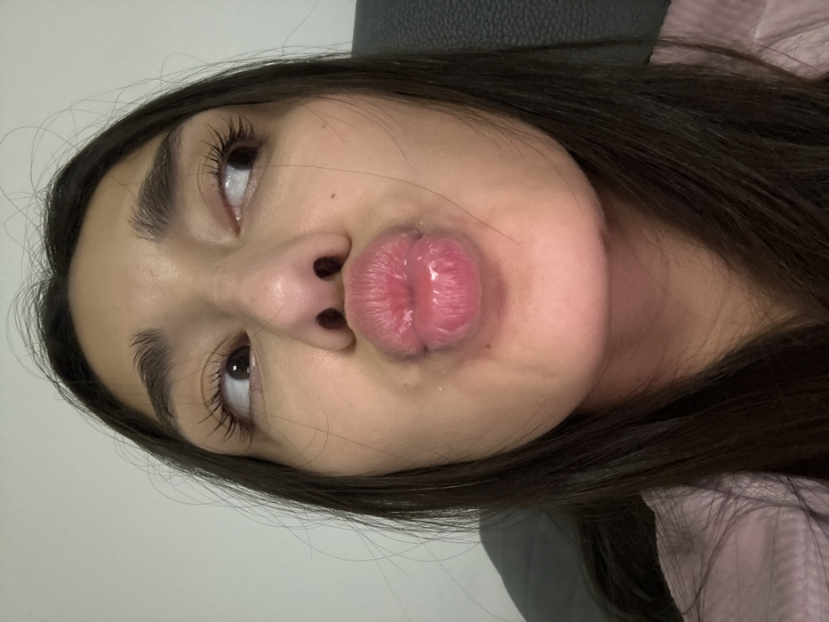 A woman's face, angled upwards, fills the frame. She purses her lips, creating a prominent pout. Her dark hair is long and partially visible. Her eyebrows are full and dark. She looks up and to the left. The background consists of a light gray wall and a hint of a gray, textured surface behind her head. She wears a light pink shirt.