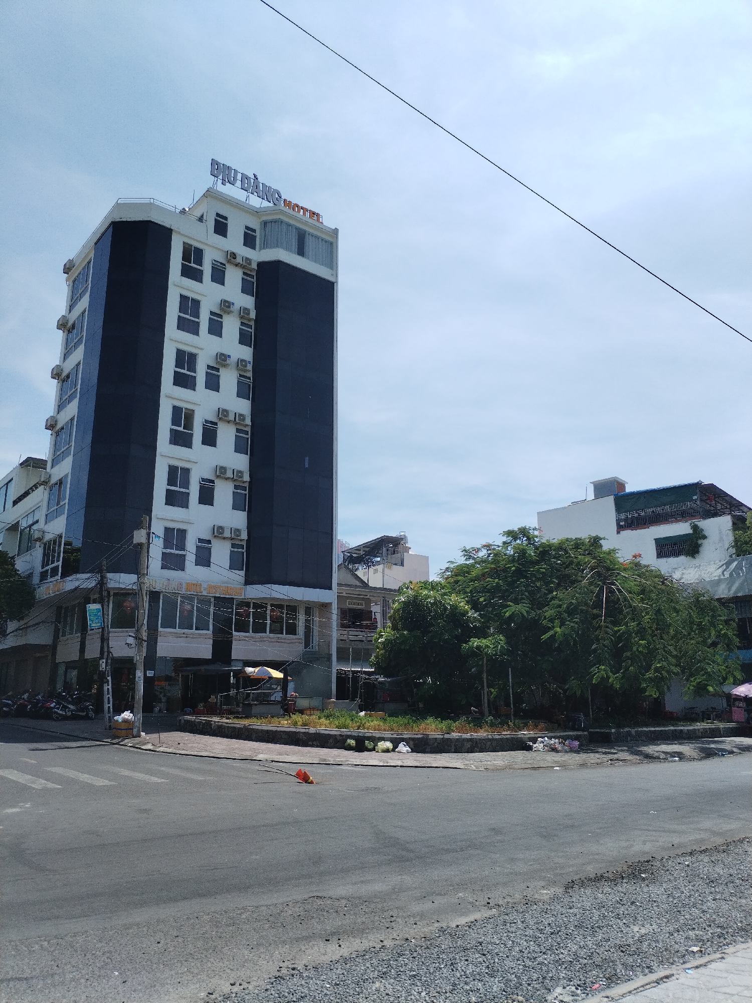 This is an outdoor, medium-angle, eye-level shot of a street intersection in Bac Nha Trang Ward, Vietnam, on a clear, bright day. A tall, modern hotel with a black and white facade dominates the left side of the image. The hotel's rooftop features blue illuminated lettering that reads "DIU DANG HOTEL." The building has multiple floors with numerous windows and external air conditioning units. Below the hotel, at street level, is a storefront with large windows. In front of the hotel, a few motorcycles are parked.
The street in the foreground is paved with asphalt and shows a pedestrian crossing. A bright orange cone lies on its side on the road near the corner. To the right of the hotel, a dense area of lush green trees obscures the view of some lower buildings. Beyond the trees, parts of other buildings are visible, including one with a green tarped roof. The environment appears to be a mix of urban development with residential and commercial structures, interspersed with natural greenery. There are no people clearly visible in the image.