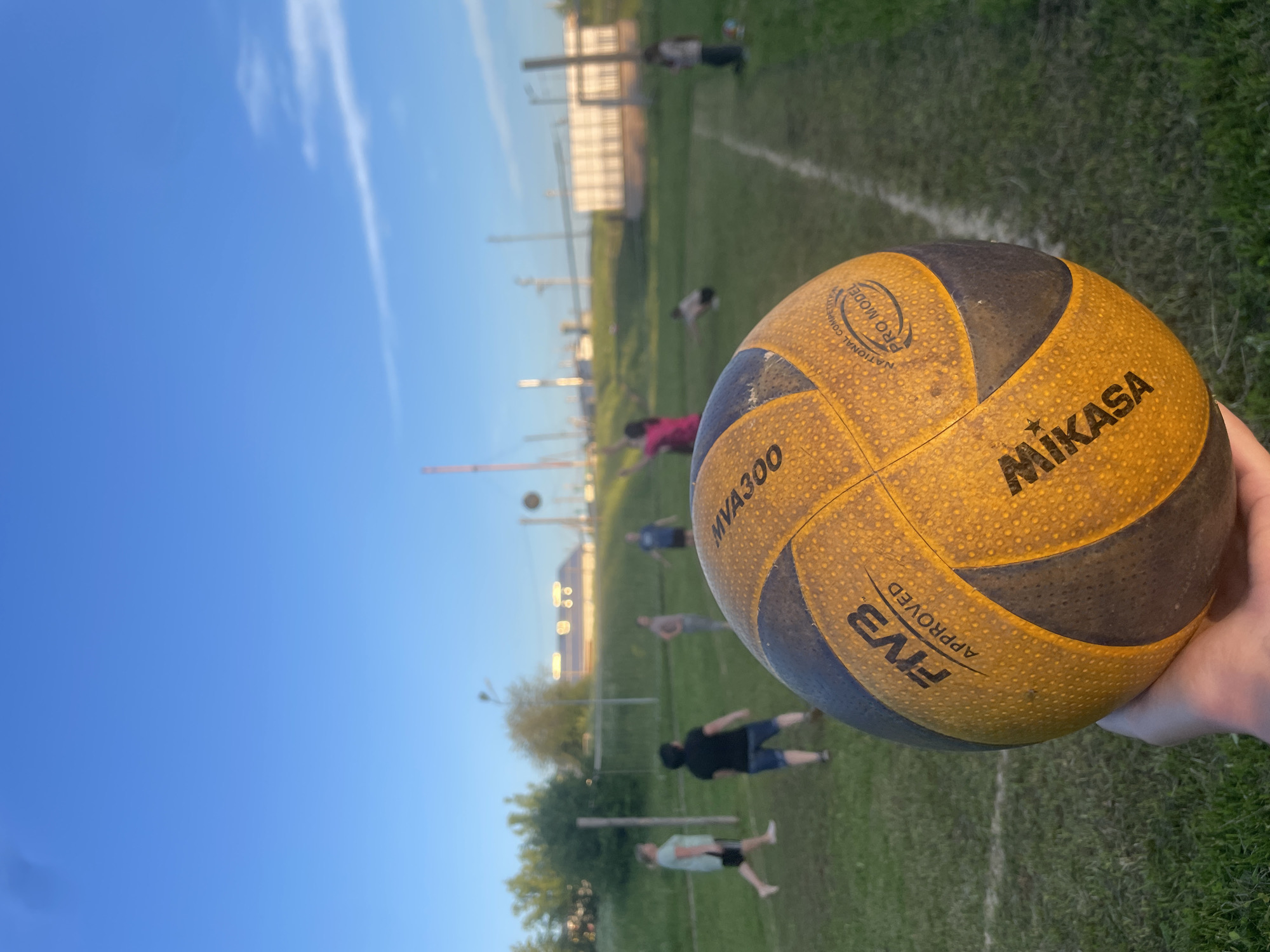 The image shows a close-up of a Mikasa volleyball held in a person's hand. The ball is yellow and black, with "FIVB APPROVED" and "MIKASA" visible. In the background, on a grassy field with white lines marking a court, a group of people are playing volleyball. The setting appears to be outdoors, with a clear blue sky and some trees and industrial buildings in the distance. The overall atmosphere suggests a casual game of volleyball. The location is identified as Podshivalovo, Russia, which is a village within the Sverdlovsk Oblast.