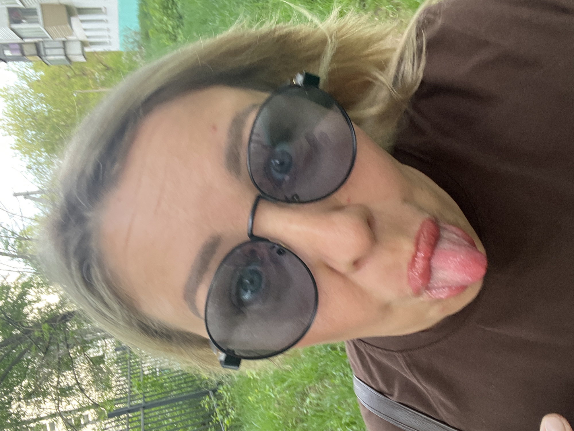 A woman is sticking out her tongue while wearing sunglasses. The background includes grass, trees, and buildings, suggesting an outdoor location in an urban area. It is impossible to verify the location is Chaikovsky, Russia, without additional information.