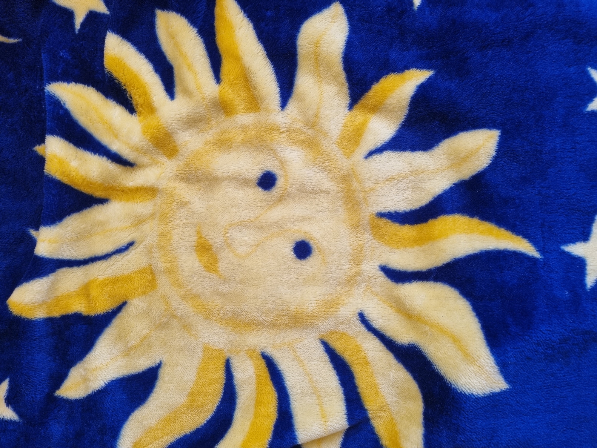 The image shows a plush blanket. The blanket has a repeating pattern of a cartoon sun with blue eyes and a smile, alternating with white stars. The blanket is primarily yellow and blue. The image does not provide any information about the location of Sochi, Russia, or any other geographic details.