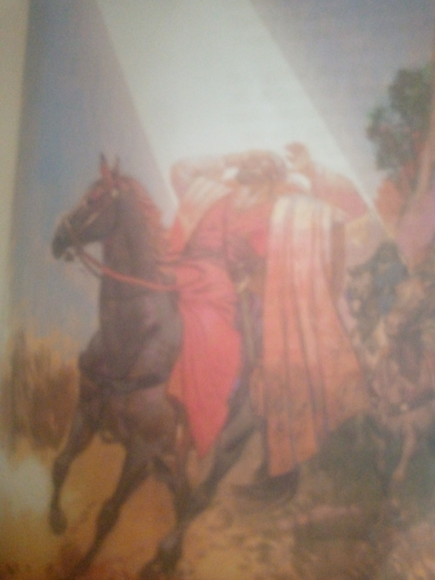 The image shows a blurred painting of a horse and rider. The horse is dark brown and appears to be in motion, possibly galloping. The rider is wearing a red garment and has their arms raised. The background is hazy and appears to be a landscape with trees and a sky illuminated by a bright light source. The overall impression is of a dynamic and dramatic scene, possibly depicting a moment of action or triumph. There is no discernible location in the city of Jauro Yinu, Nigeria, within the painting itself.