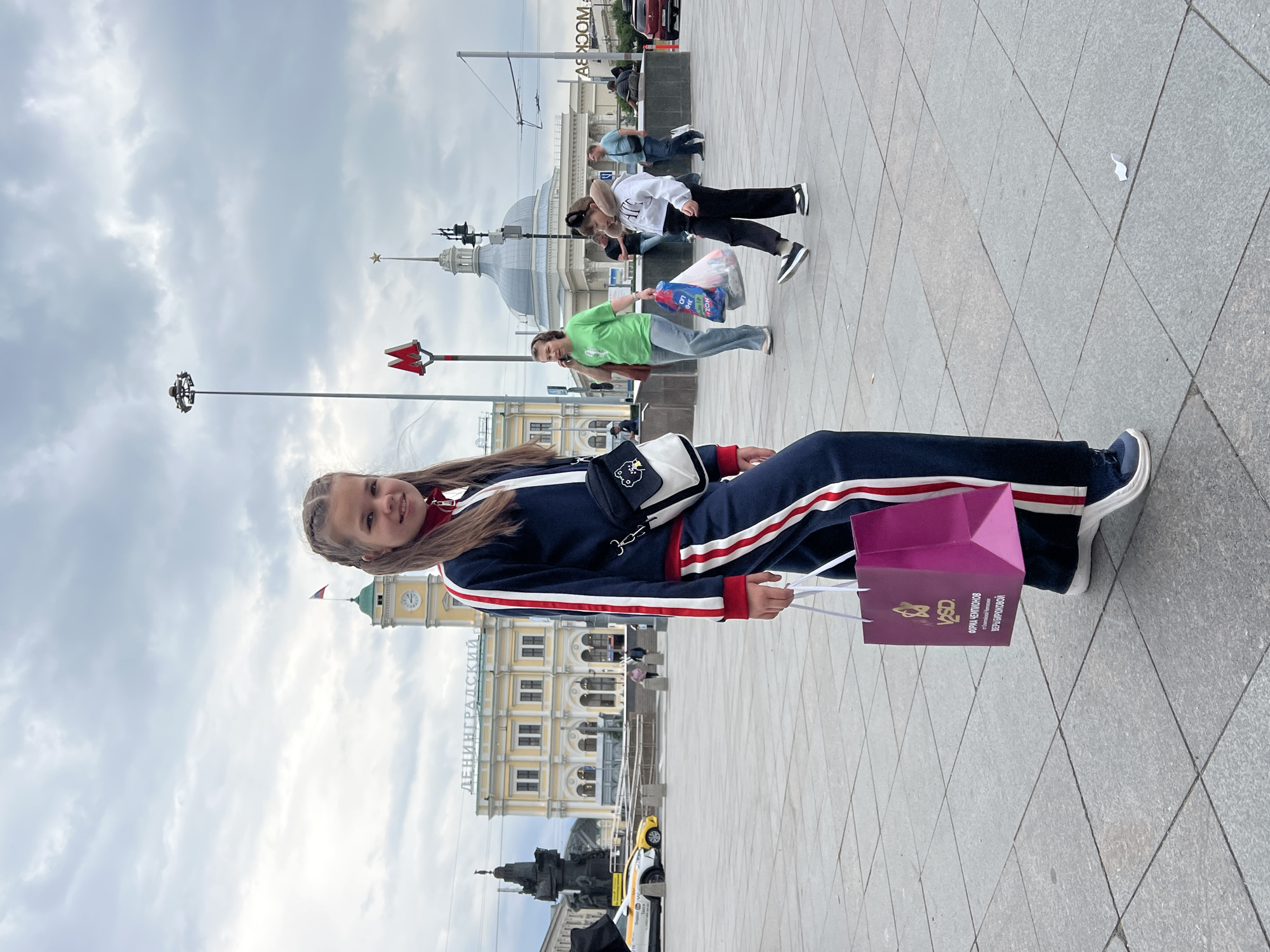 A young girl with braided hair is standing in a paved plaza in front of a yellow building with a clock tower. She is wearing a navy blue tracksuit with red and white stripes and carrying a pink shopping bag and a small white crossbody bag. In the background, other people are walking, and there are several buildings, including a domed structure and a metro sign. The sky is overcast. This scene is likely Leningradsky Rail Terminal Square in Moscow, Russia, given the architectural style and the presence of the Leningradsky station building.