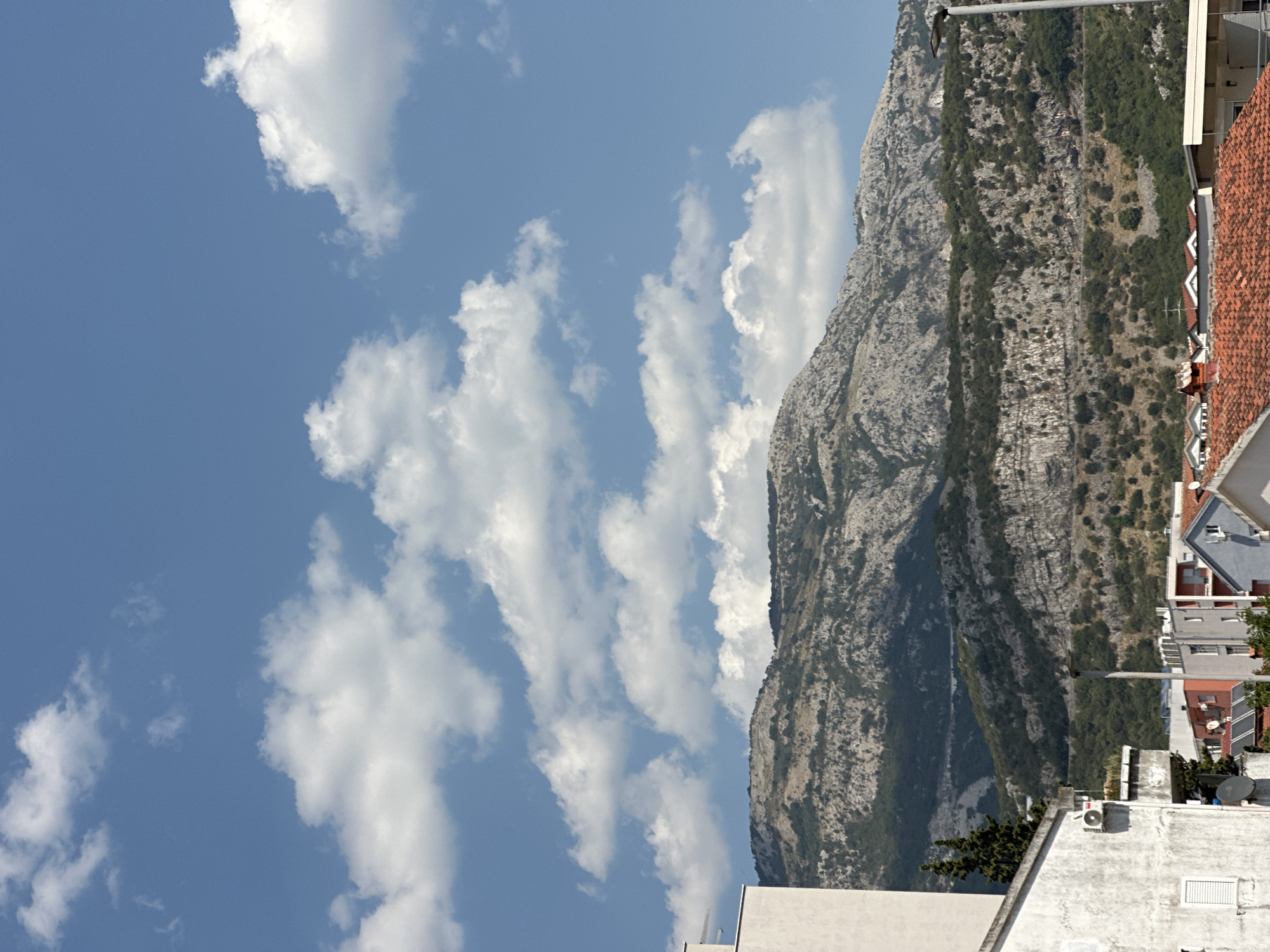 The image captures a daytime scene with a vast blue sky filled with fluffy white clouds. In the middle ground, a rugged, rocky mountain range dominates the landscape, its slopes covered in patches of green vegetation. Below the mountains, a cluster of buildings with red tile roofs and white or light-colored facades forms a residential area. The foreground features a portion of a light-colored building, partially visible on the left side of the frame. A street lamp is visible on the right, extending upwards towards the sky. The environment suggests a Mediterranean setting, possibly the city of Budva, Montenegro, known for its mountainous backdrop and coastal location. There are no people or animals visible in the image.