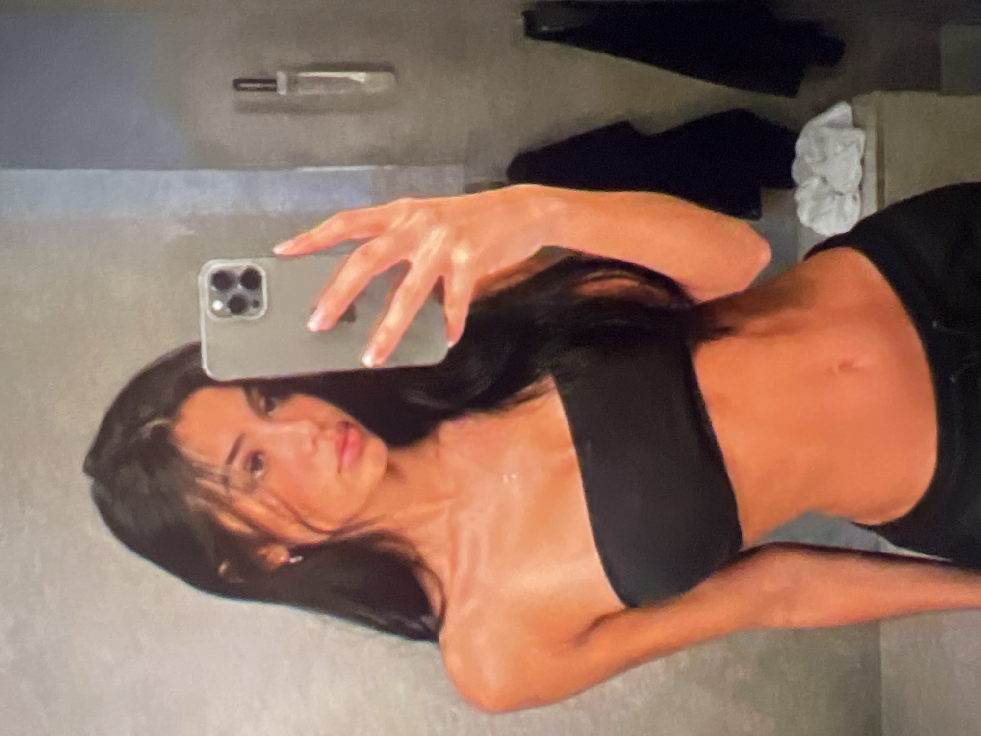 The image is a selfie of a young woman with long, dark hair, wearing a black strapless top and dark pants. She is holding a smartphone in her right hand, seemingly taking the photo. The background appears to be a bathroom with a gray textured wall. A light-colored shower curtain is visible on the right, and a white folded cloth is placed on a surface behind her. The lighting is warm and casts a glow on her skin. The location is unknown.