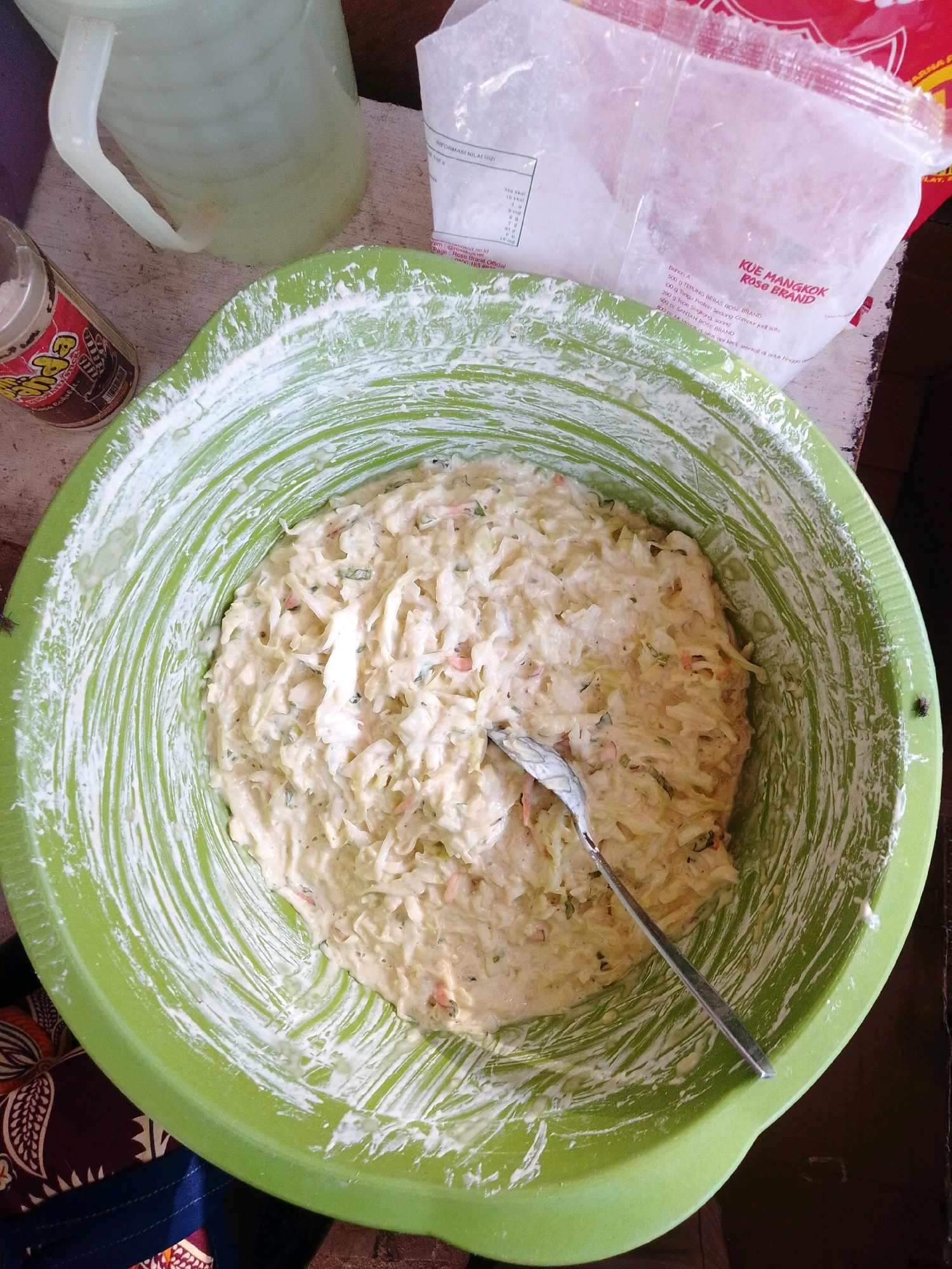 A close-up, high-angle shot shows a large green plastic bowl filled with a pale, creamy batter, likely for a cake or fritter. The batter appears to contain shredded vegetables, possibly cabbage and carrots, and flecks of herbs or spices. The inside of the bowl is splattered with a white substance, likely flour, which has been mixed into the batter. A silver spoon rests in the batter, angled towards the right. To the left of the bowl, a glass pitcher with a handle is partially visible, containing a clear liquid. In the background, a white plastic bag with Indonesian text is draped over a surface, possibly a table or counter. The overall scene suggests food preparation. The presence of Indonesian text on the packaging indicates the location is likely Indonesia. While the specific city of Cikal is mentioned, there are no visual elements within the image that definitively confirm this location.