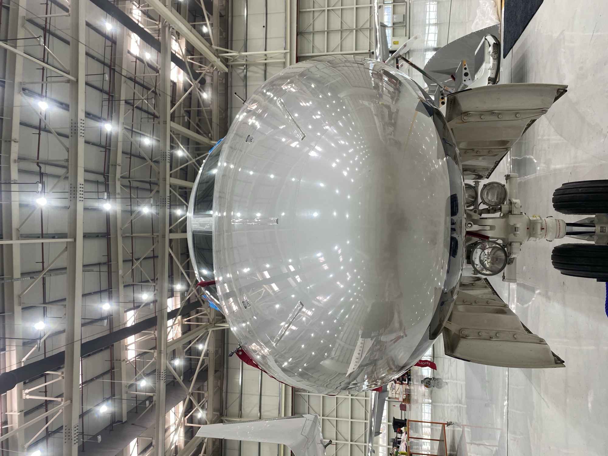 The image captures the front section of a large, highly polished silver aircraft, likely a commercial or business jet, situated indoors within a spacious aircraft hangar or maintenance facility. The main subject is the rounded nose cone and cockpit windows of the aircraft, which intensely reflect the bright overhead artificial lighting and the structural elements of the hangar.
The aircraft's nose landing gear is partially visible on the right side of the frame, featuring dual wheels, a strut, and a prominent landing light. Near the top of the nose, a pitot tube can be seen protruding. The polished surface of the fuselage reflects numerous bright rectangular and circular light sources, creating a scattered, dazzling pattern. Part of a wing or horizontal stabilizer is also visible in the lower left, indicating the scale of the aircraft.
The background reveals the vast interior of the hangar, characterized by a high ceiling supported by an intricate network of white and beige structural beams, girders, and trusses. Red piping runs along some of the vertical supports. Numerous bright lights are mounted across the ceiling and structural framework, illuminating the entire space. The floor is a light, clean surface, also contributing to the reflections on the aircraft.
The aircraft appears to be stationary, suggesting it is undergoing maintenance, inspection, or storage within the hangar. The scene conveys an industrial and technical environment. No people are explicitly visible. Faint, distorted alphanumeric characters, possibly "MIO-XL" or similar, are reflected on the aircraft's lower fuselage. Additionally, text on the landing gear appears to include "LIGHT". The location context provided indicates this scene is in Dubai, United Arab Emirates.