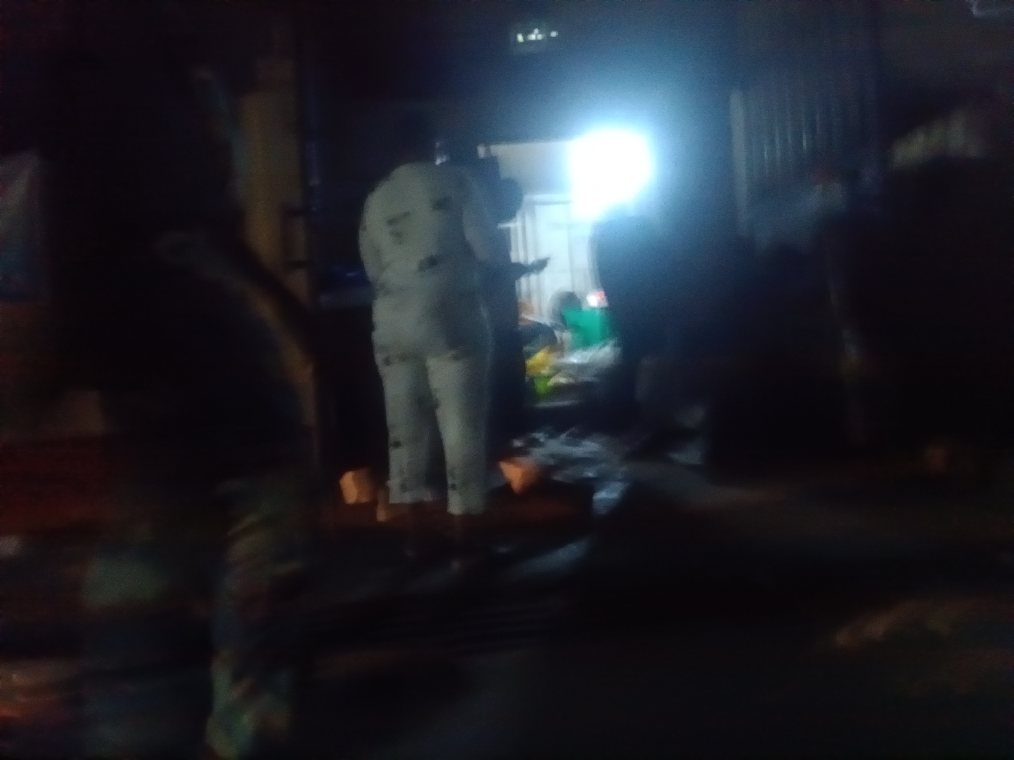 The image depicts a dimly lit scene, possibly at night, within a commercial setting in Ado, Nigeria. In the foreground, a person is visible from the waist down, wearing light-colored pants with a patterned design and dark footwear. Their stance suggests they are standing still. To the left, the silhouette of another person or object is partially visible. In the midground, another individual stands facing away from the camera, wearing a light-colored garment, possibly a uniform or tracksuit, with dark markings. This person is holding a dark object, perhaps a phone or a small device, in their hands. Behind them, a bright light source illuminates the area, creating a glare. Scattered objects, some appearing to be light-colored and possibly containers or blocks, are visible on the ground in the midground. The background is dark and indistinct, with vertical lines suggesting a structure like a corrugated iron fence or stall. The overall impression is of an active, albeit somewhat chaotic, environment.